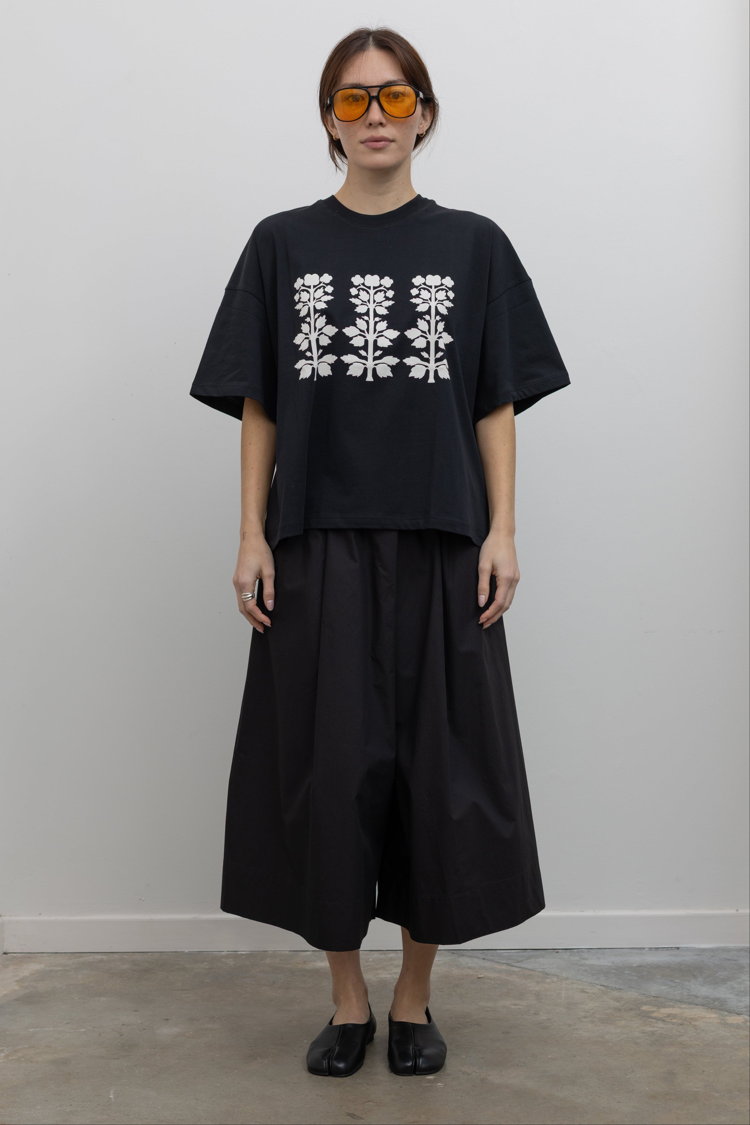 Full front view of a woman standing wearing spring fashion in a Kowtow black cotton jersey relaxed-fit short-sleeve tee featuring a contrasted screen-printed floral design on the front at Alas Eius.