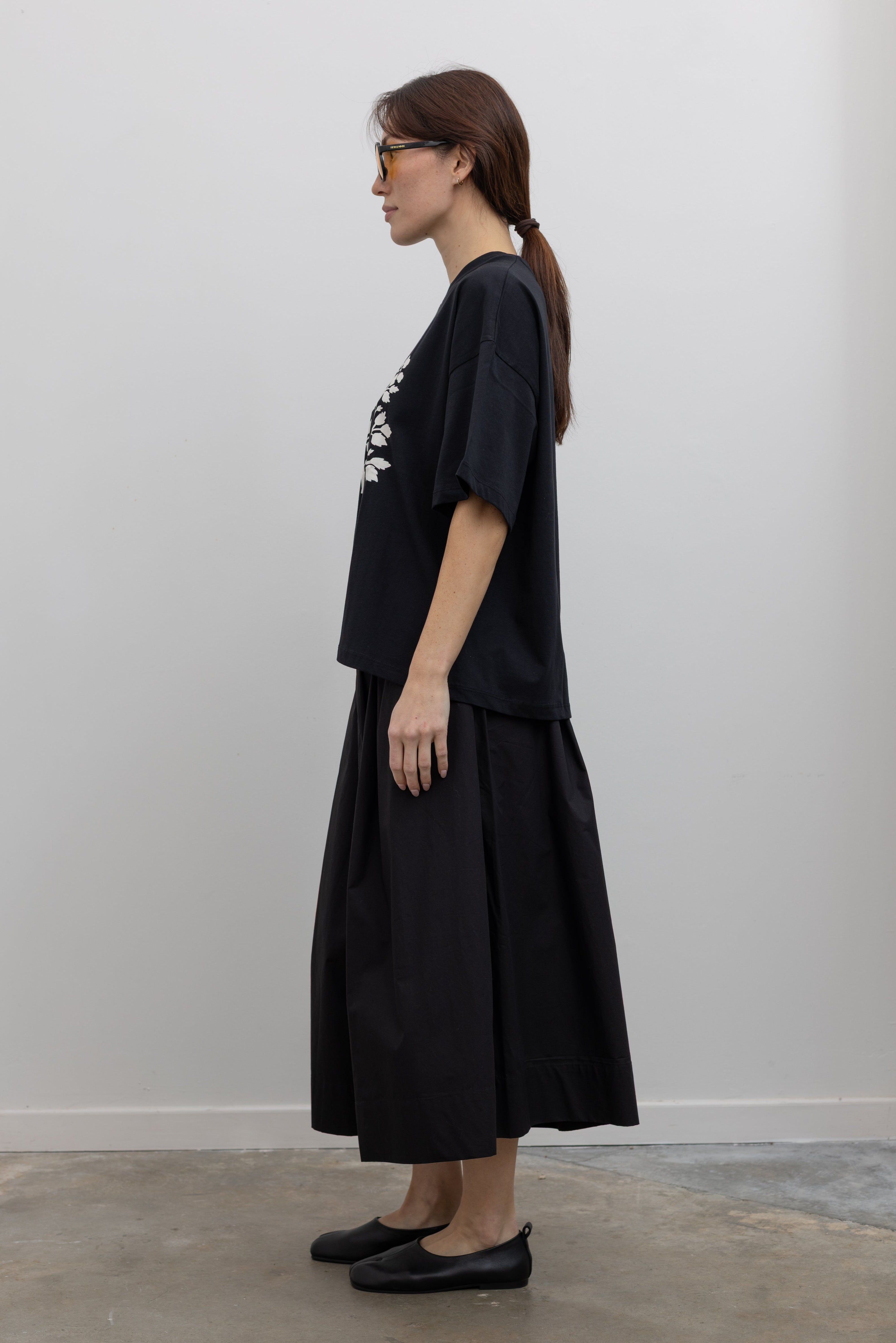 Full side view of a woman standing wearing spring fashion in a Kowtow black cotton jersey relaxed-fit short-sleeve tee featuring a contrasted screen-printed floral design on the front at Alas Eius.