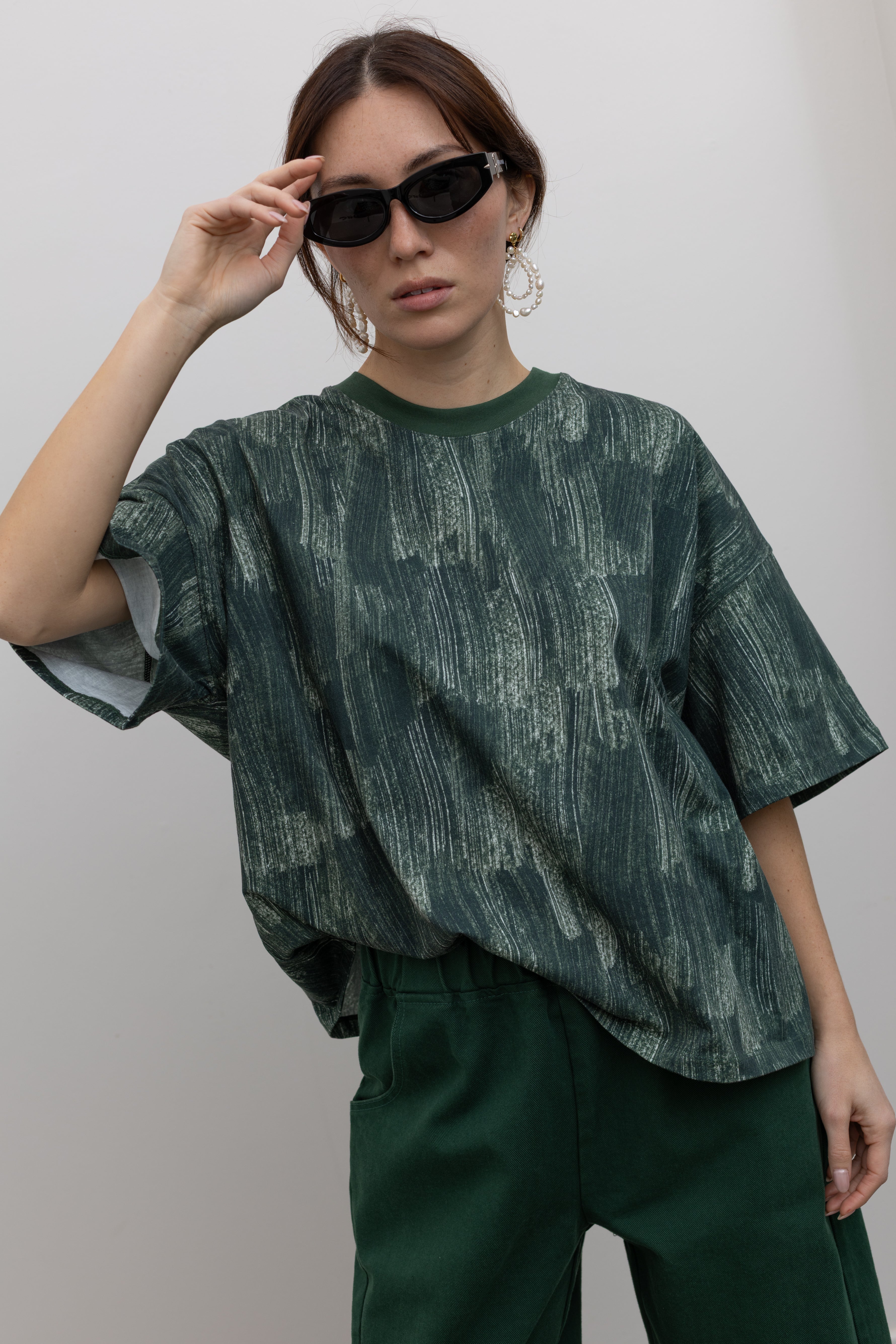 Close-up front view of a woman posing with sunglasses wearing spring fashion in a Kowtow green oversized short sleeve t-shirt with an all over white brushstroke print paired with pine colored denim jeans at Alas Eius.