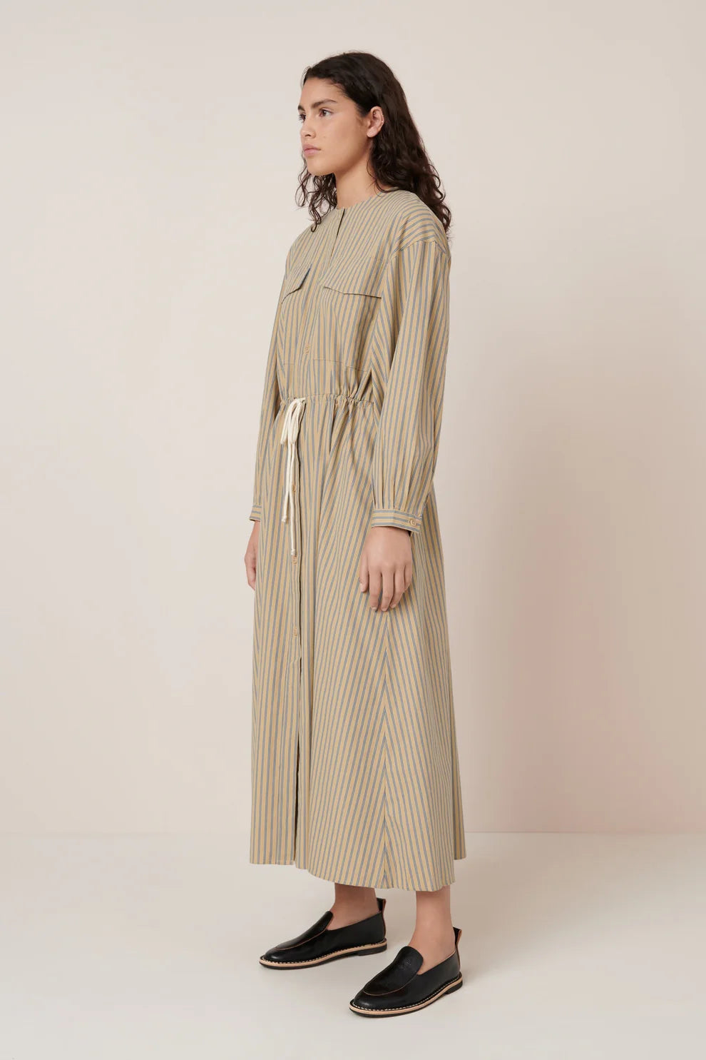 Full 3/4 view of a woman standing wearing Spring fashion in a Kowtow Array double pinstripe fairtrade organic cotton poplin dress featuring oversized front pockets, a slightly dropped drawcord waist, and fastened with nut buttons down the front at Alas Eius.
