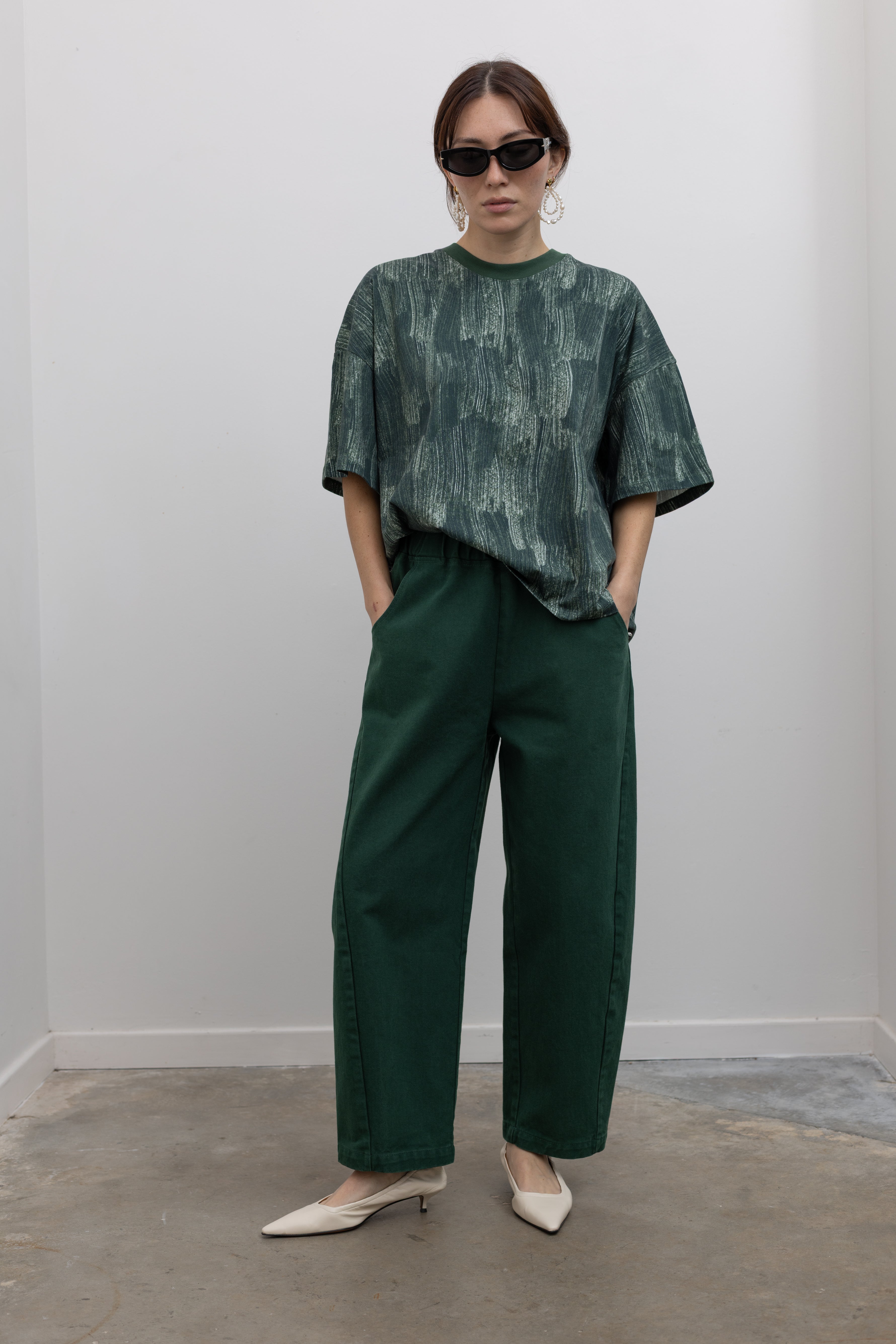 Front full view of a woman posing with hands in jean pockets wearing spring fashion in a Kowtow green oversized short sleeve t-shirt with an all over white brushstroke print paired with pine colored denim jeans at Alas Eius.