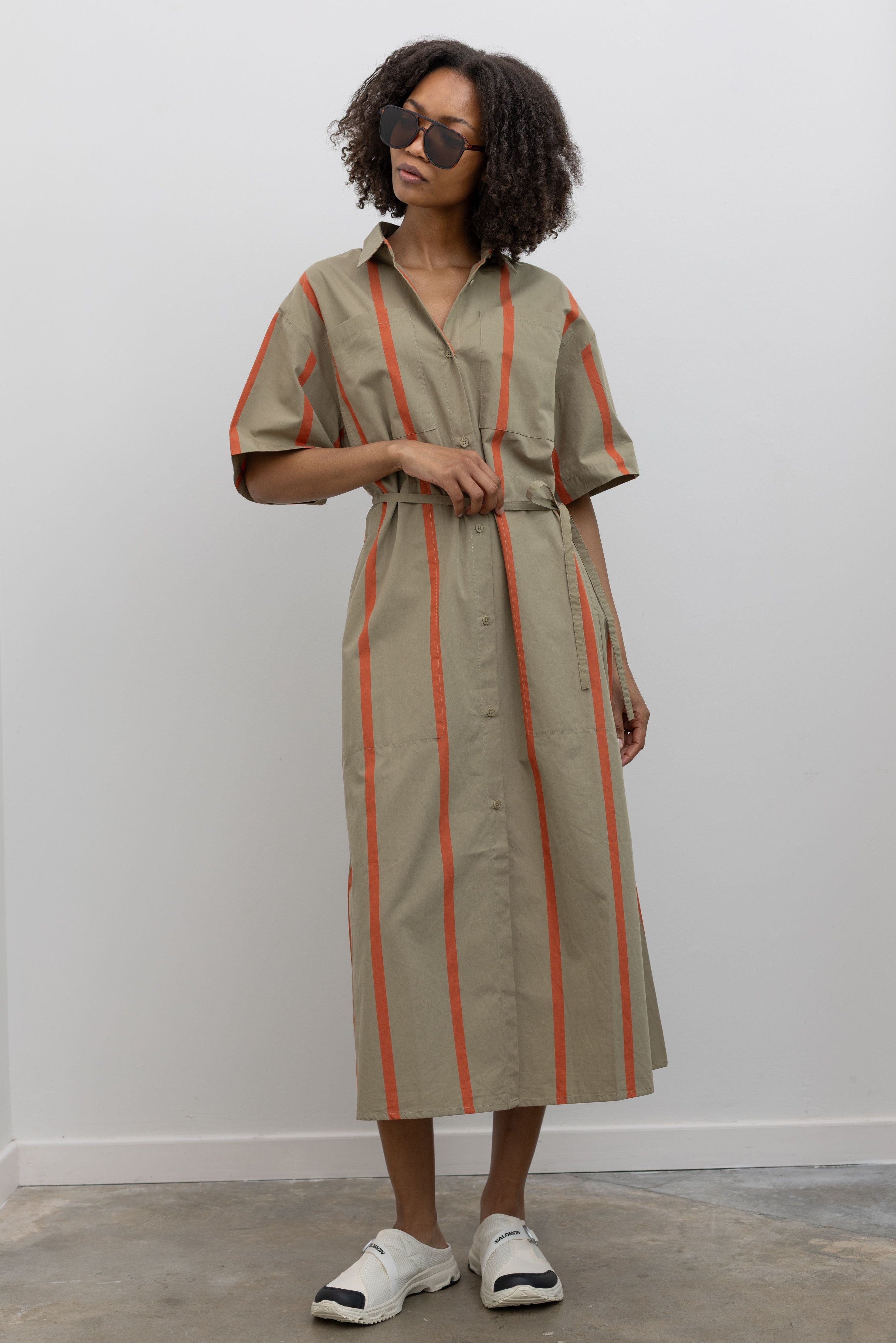 Full length front view of a woman posing wearing elevated casual weekend spring fashion in a Kowtow Organic Cotton Nasturtium Stripe Oversized Balance Dress featuring pockets, a classic collar, nut buttons down the front, and a tie belt paired with Salomon slip on sneakers at Alas Eius. 