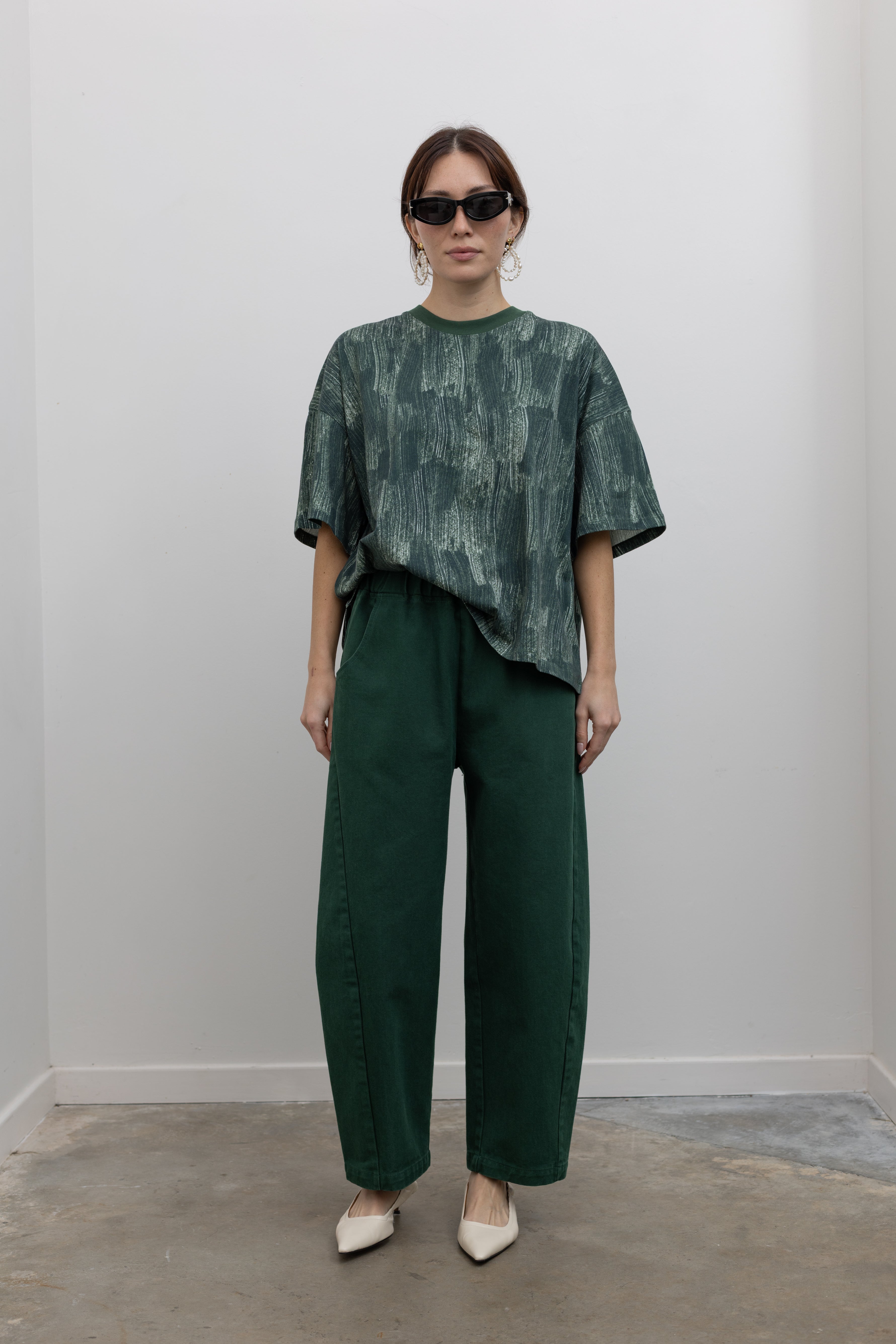 Full front view of a woman posing with sunglasses wearing spring fashion in a Kowtow green oversized short sleeve t-shirt with an all over white brushstroke print paired with pine colored denim jeans at Alas Eius.