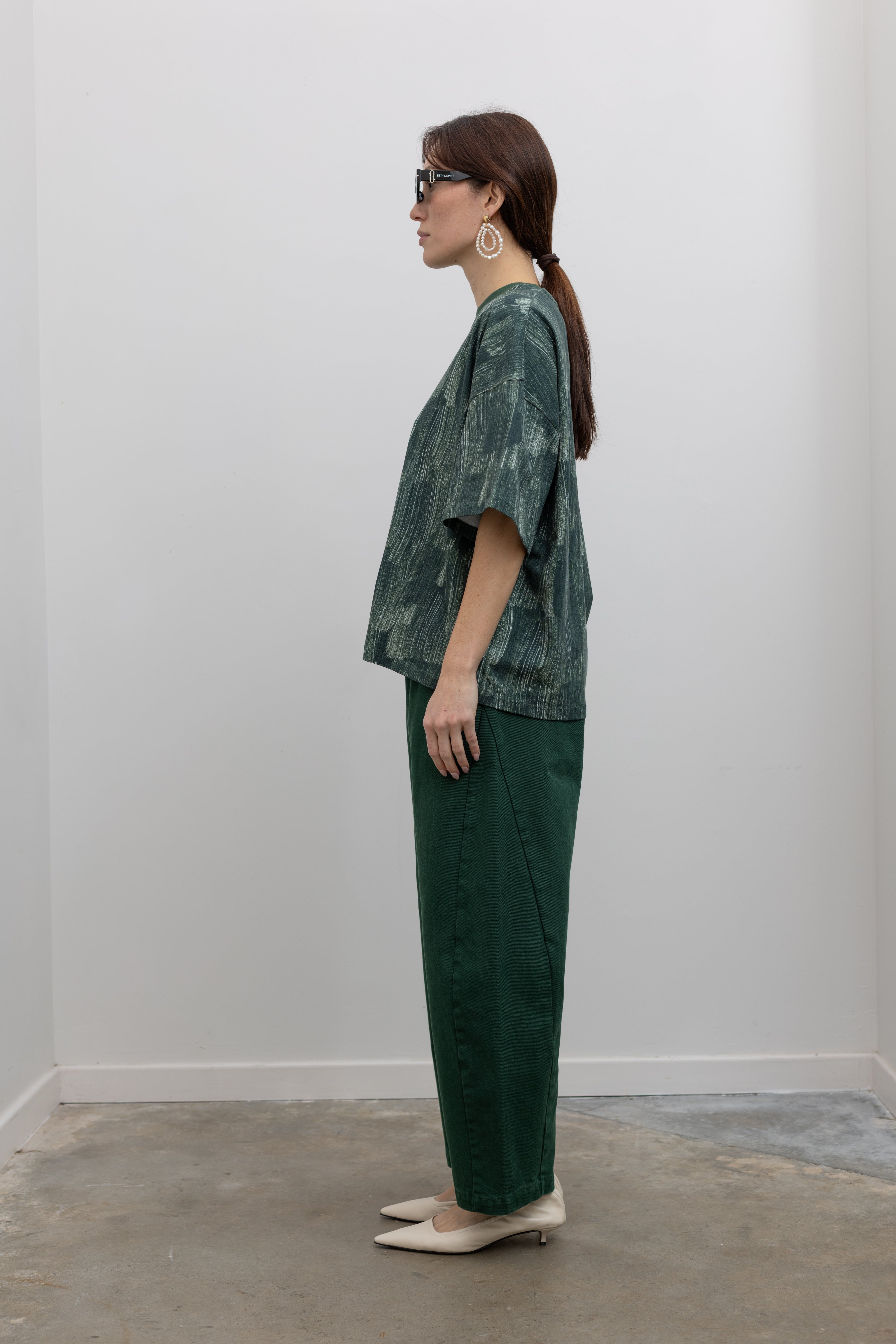 Full side view of a woman posing with sunglasses wearing spring fashion in a Kowtow green oversized short sleeve t-shirt with an all over white brushstroke print paired with pine colored denim jeans at Alas Eius.