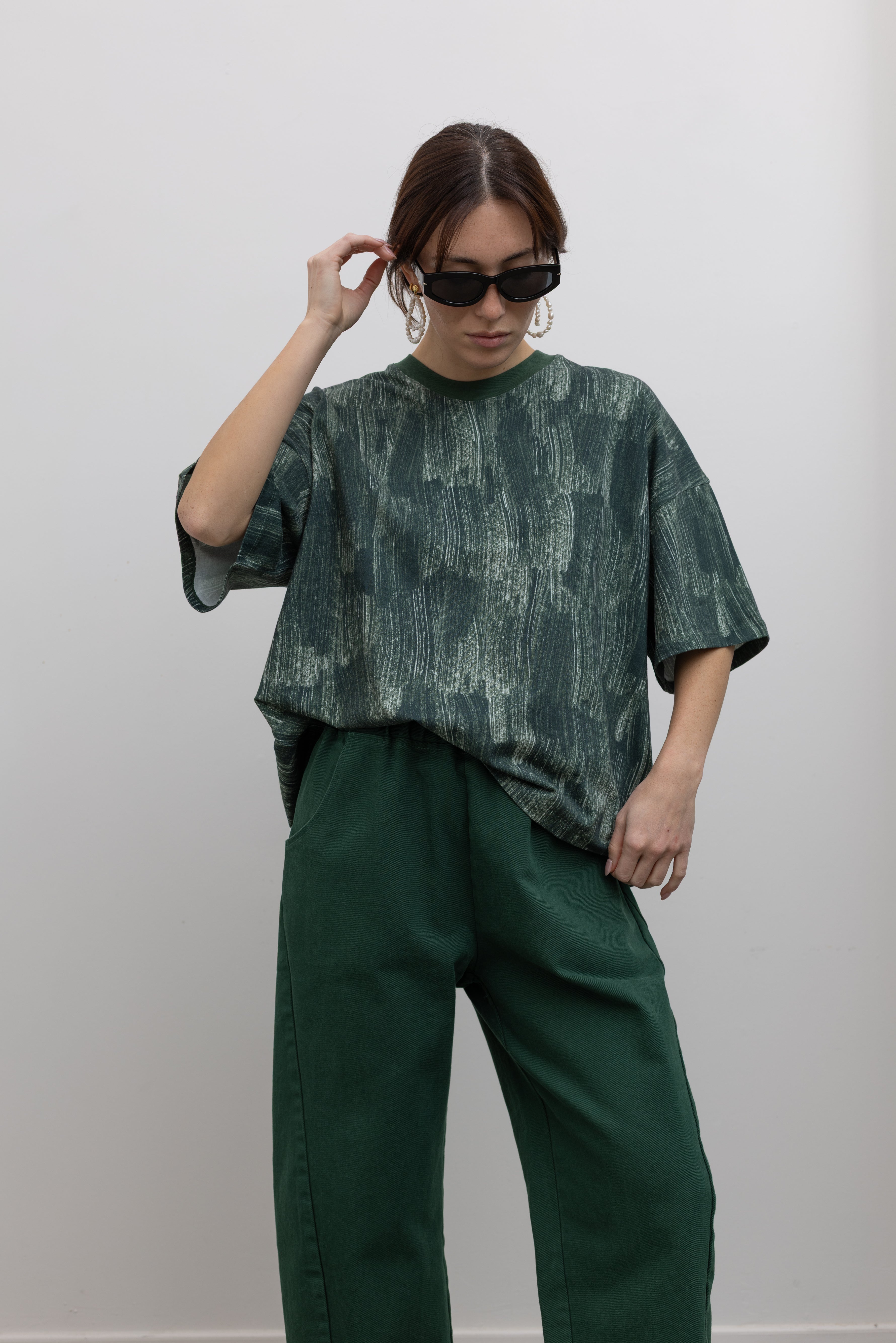 Front view of a woman posing with sunglasses wearing spring fashion in a Kowtow green oversized short sleeve t-shirt with an all over white brushstroke print paired with pine colored denim jeans at Alas Eius.