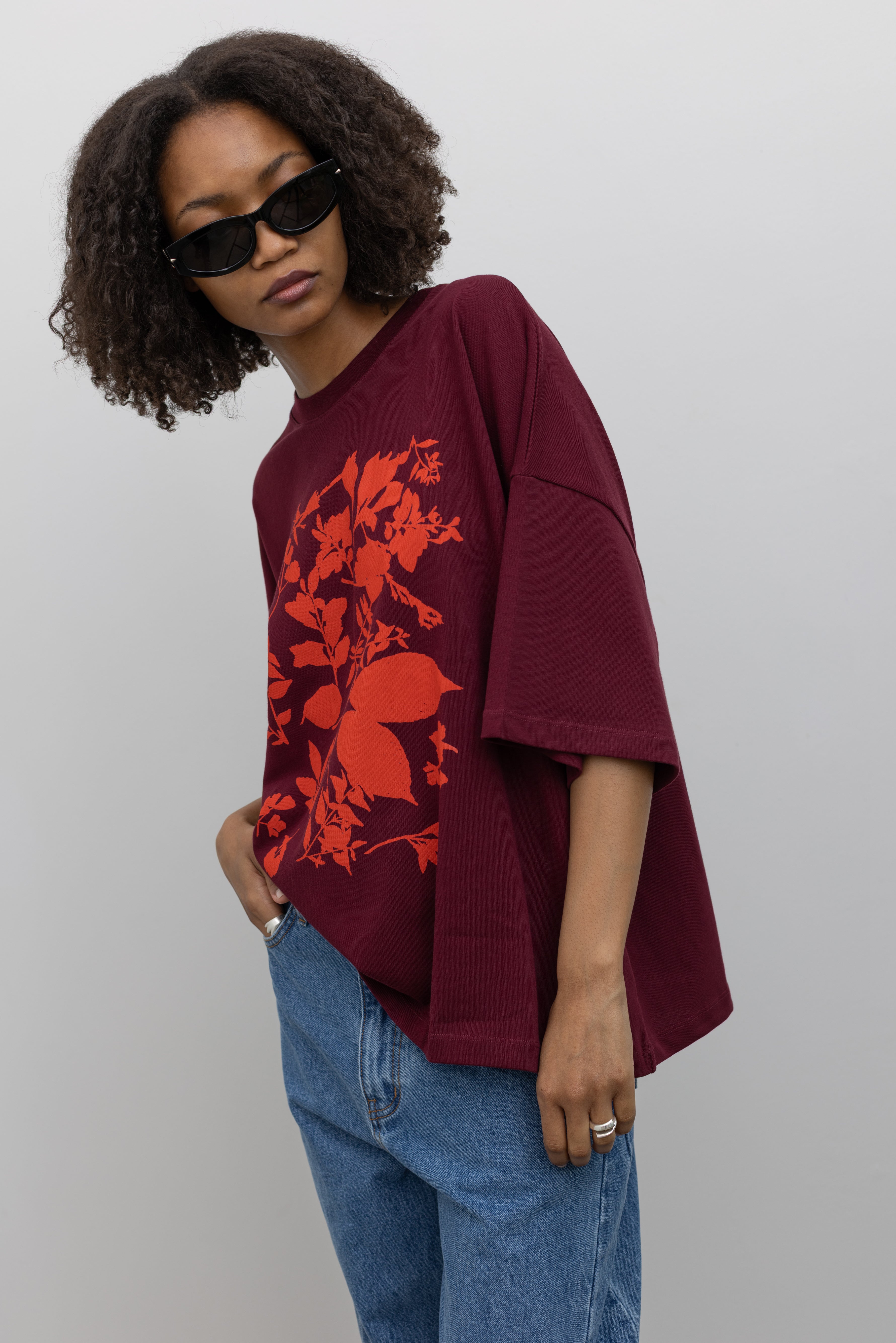 Full length back view of a woman posing wearing an elevated summer staple in a Kowtow Maroon Stencil Tee featuring a screen-printed pressed foliage motif with short sleeves, an oversized fit, and a ribbed crew neck paired with light wash relaxed fit denim jeans at an Alas Eius store. 