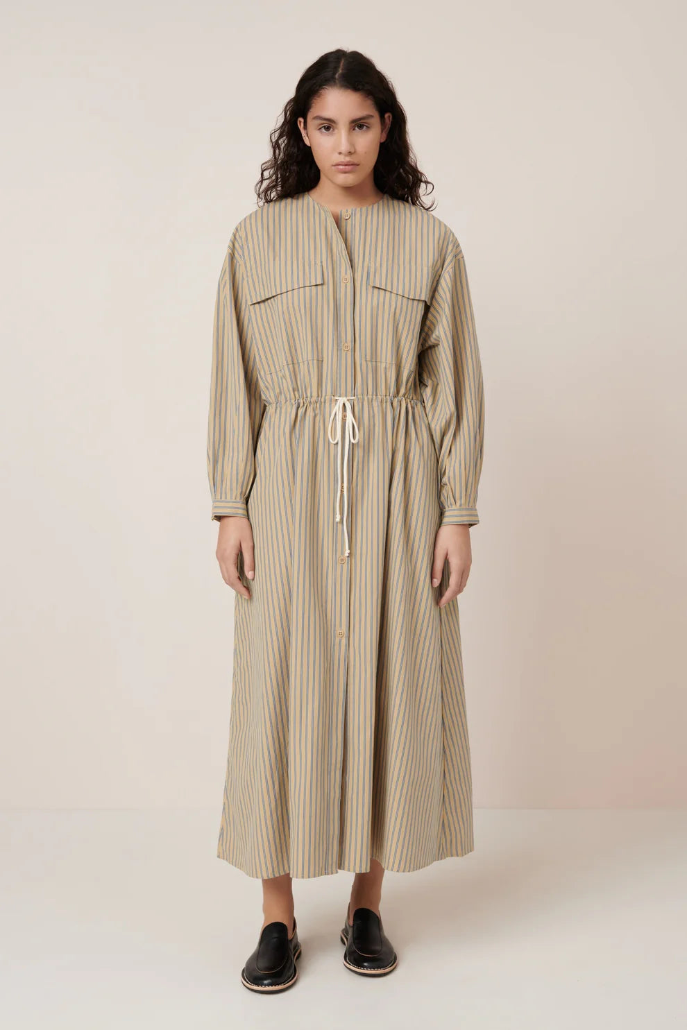 Full front view of a woman standing wearing Spring fashion in a Kowtow Array double pinstripe fairtrade organic cotton poplin dress featuring oversized front pockets, a slightly dropped drawcord waist, and fastened with nut buttons down the front at Alas Eius.