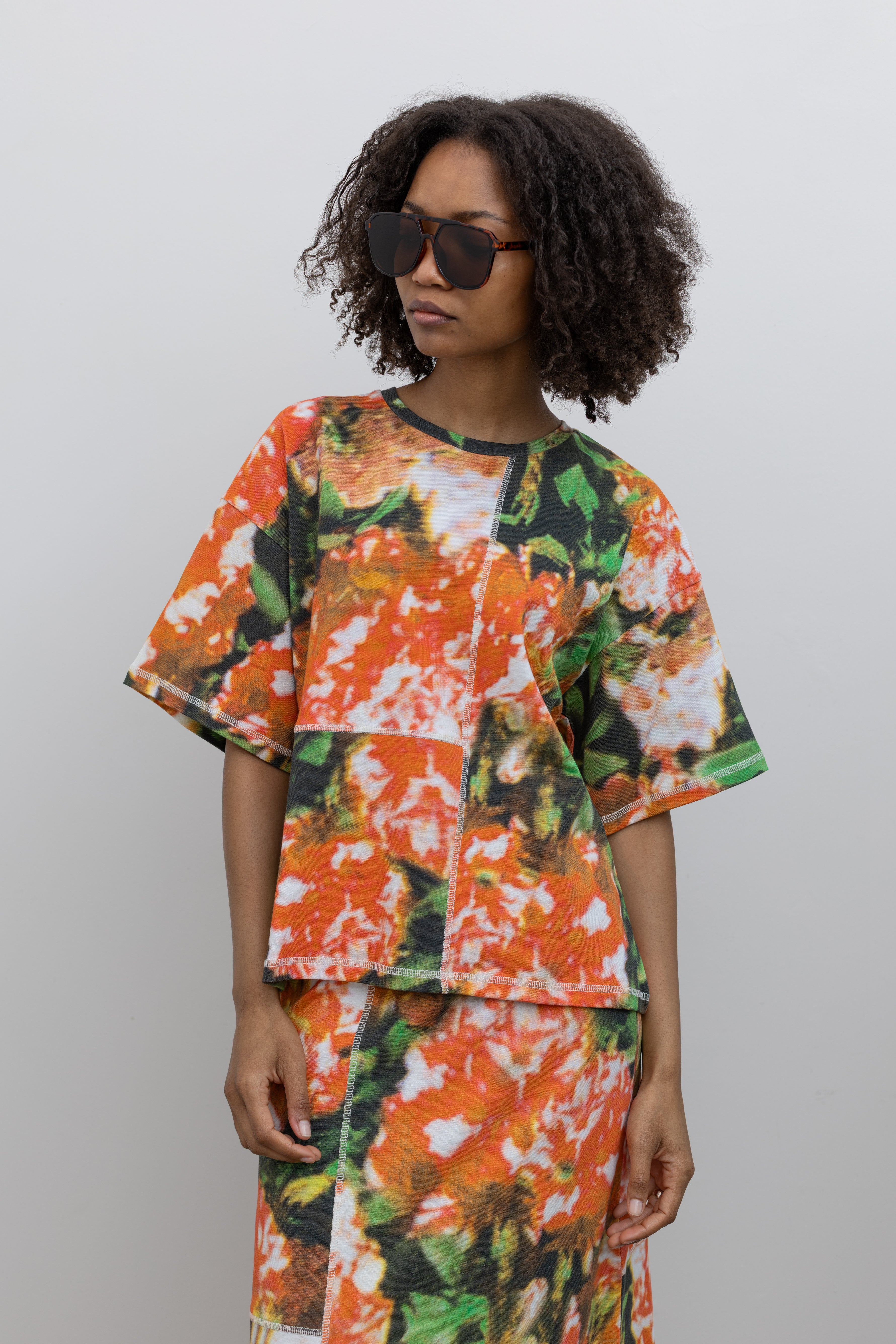 Close-up front view of a woman wearing Spring and Summer fashion in a vibrant and abstract print Kowtow Flower Book Montage Tee featuring a boxy shape, drop shoulders, and patchwork panelling throughout that is highlighted with contrast white top stitching paired with its matching skirt to create the perfect weekend outfit at Alas Eius. 