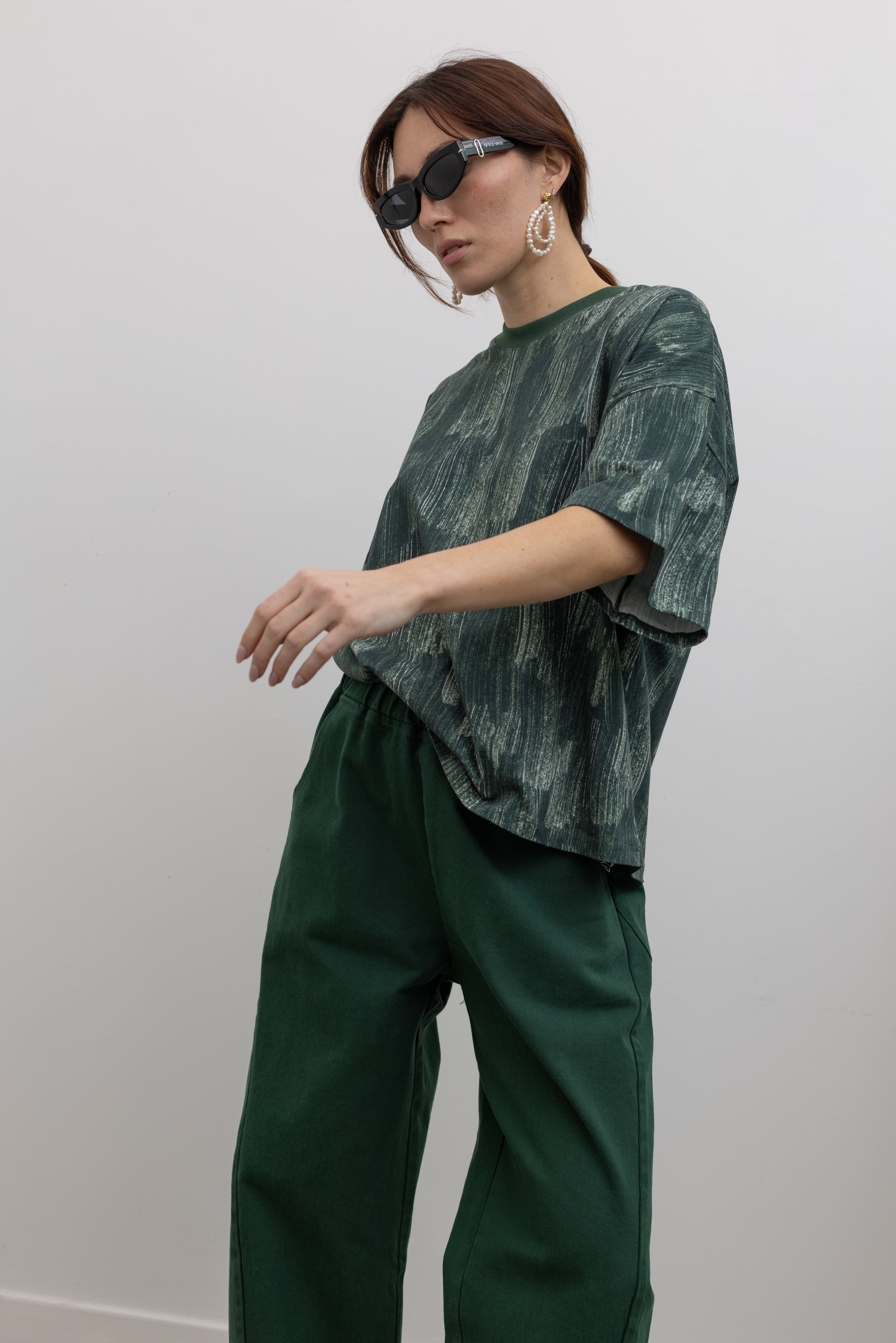 Front view of a woman in motion with wearing spring fashion in a Kowtow green oversized short sleeve t-shirt with an all over white brushstroke print paired with pine colored denim jeans at Alas Eius.