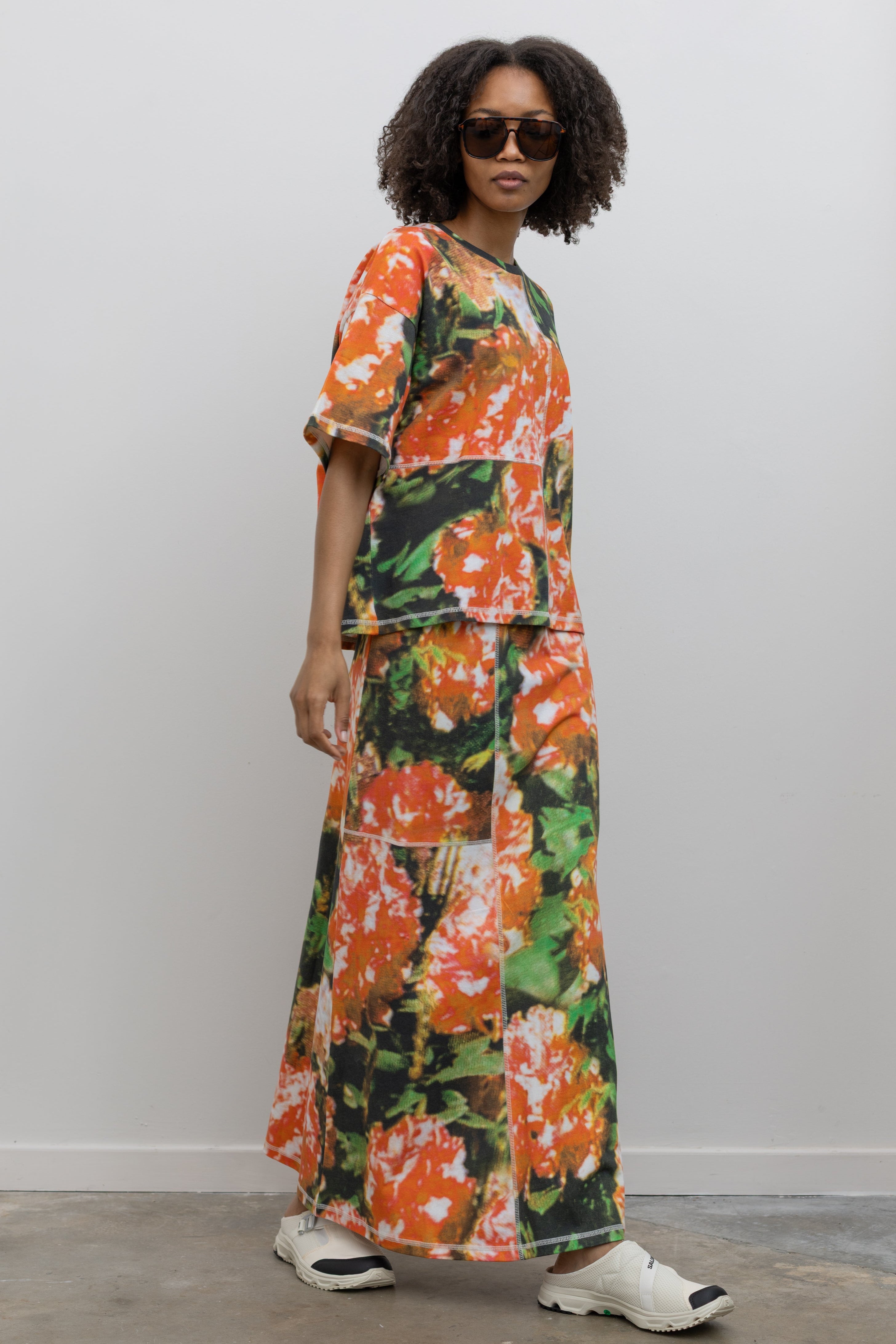 Full length front view of a woman wearing an elevated set in a vibrant and abstract print Kowtow Flower Book Montage Tee featuring a boxy shape, drop shoulders, and patchwork panelling throughout that is highlighted with contrast white top stitching paired with the Flower Book Montage maxi skirt at an Alas Eius store. 