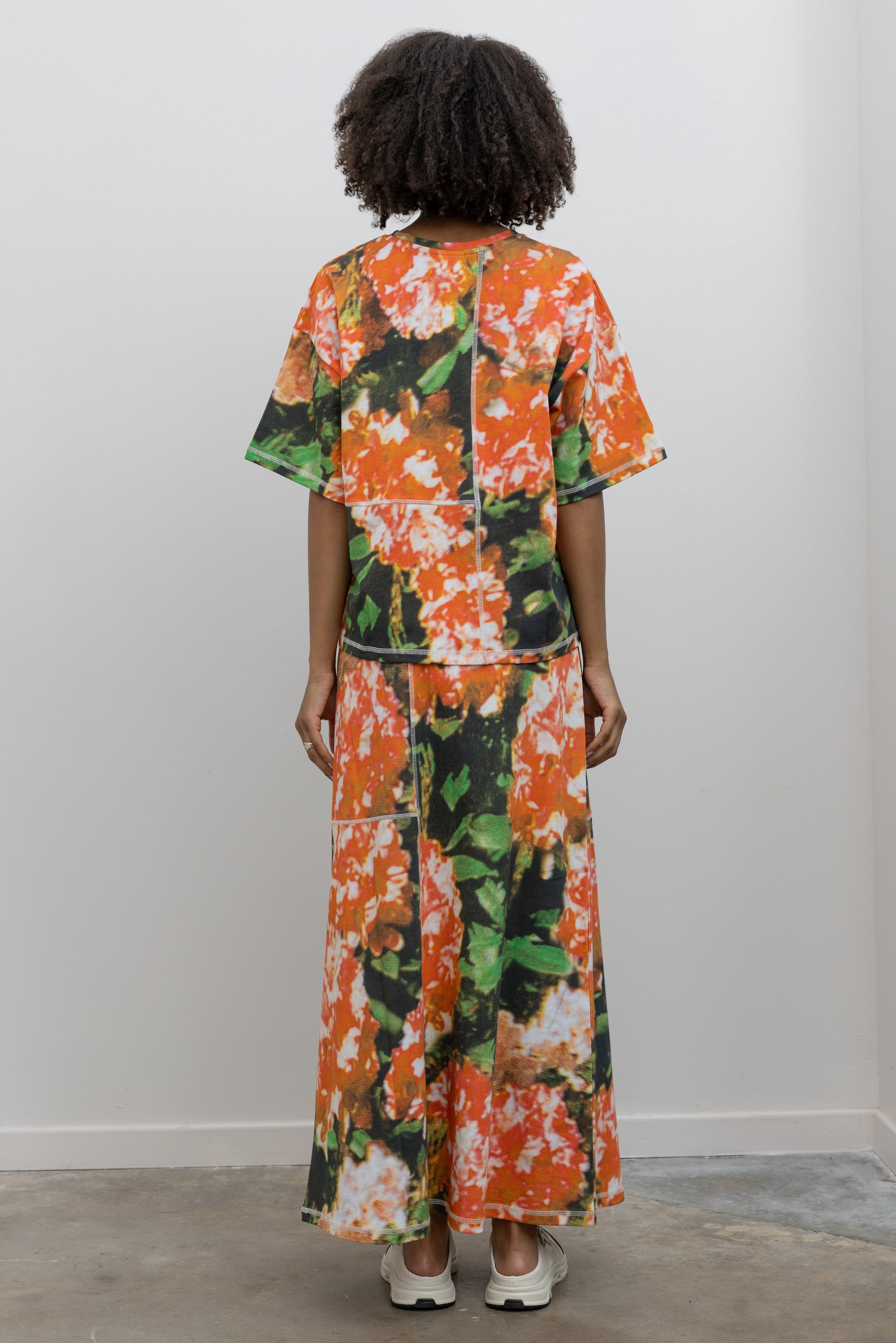 Full length back view of a woman wearing a summer coordinating set in a vibrant and abstract print Kowtow Flower Book Montage Tee featuring a boxy shape, drop shoulders, and patchwork panelling throughout that is highlighted with contrast white top stitching paired with its matching Flower Book Montage maxi skirt at an Alas Eius store. 