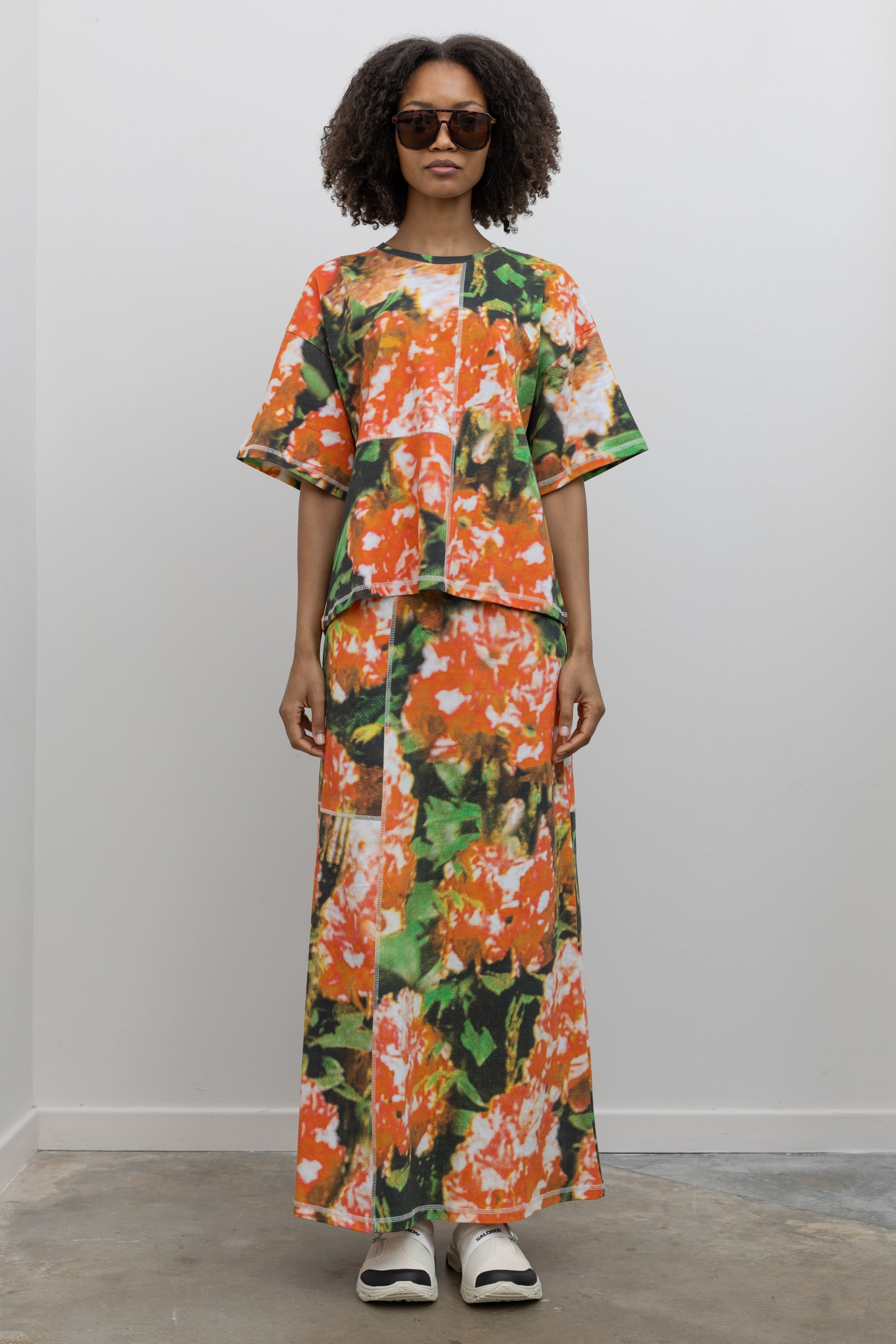 Full length front view of a woman wearing the perfect travel set in a vibrant and abstract print Kowtow Flower Book Montage Tee featuring a boxy shape, drop shoulders, and patchwork panelling throughout that is highlighted with contrast white top stitching paired with its matching Flower Book Montage maxi skirt at an Alas Eius store. 