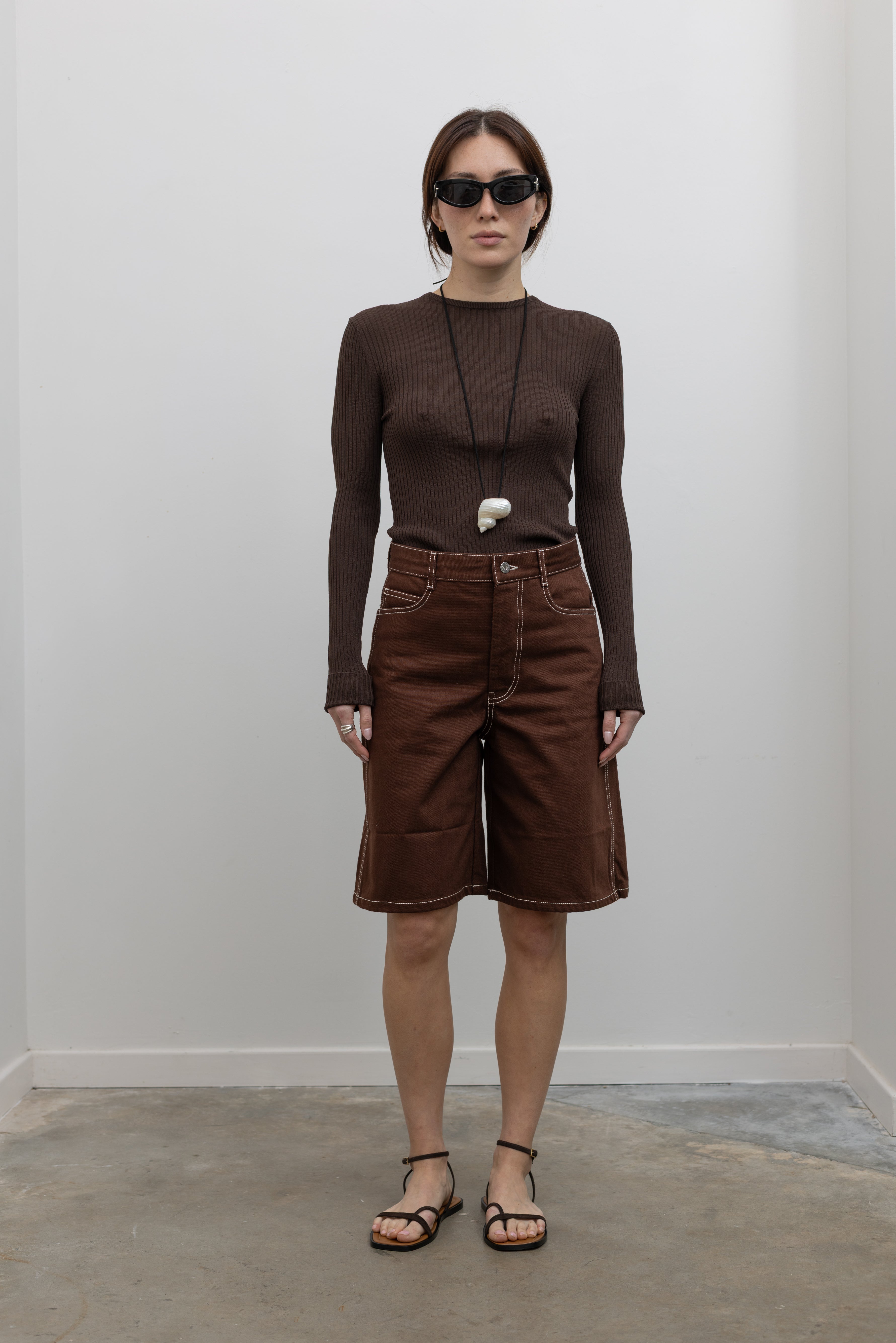 Woman wearing Spring fashion in a pair of Kowtow Framework Brown relaxed, straight fit Organic Cotton Denim Shorts with contrast white topstitching and brown long-sleeved ribbed top with a shell necklace, sunglasses, and strappy sandals at Alas Eius