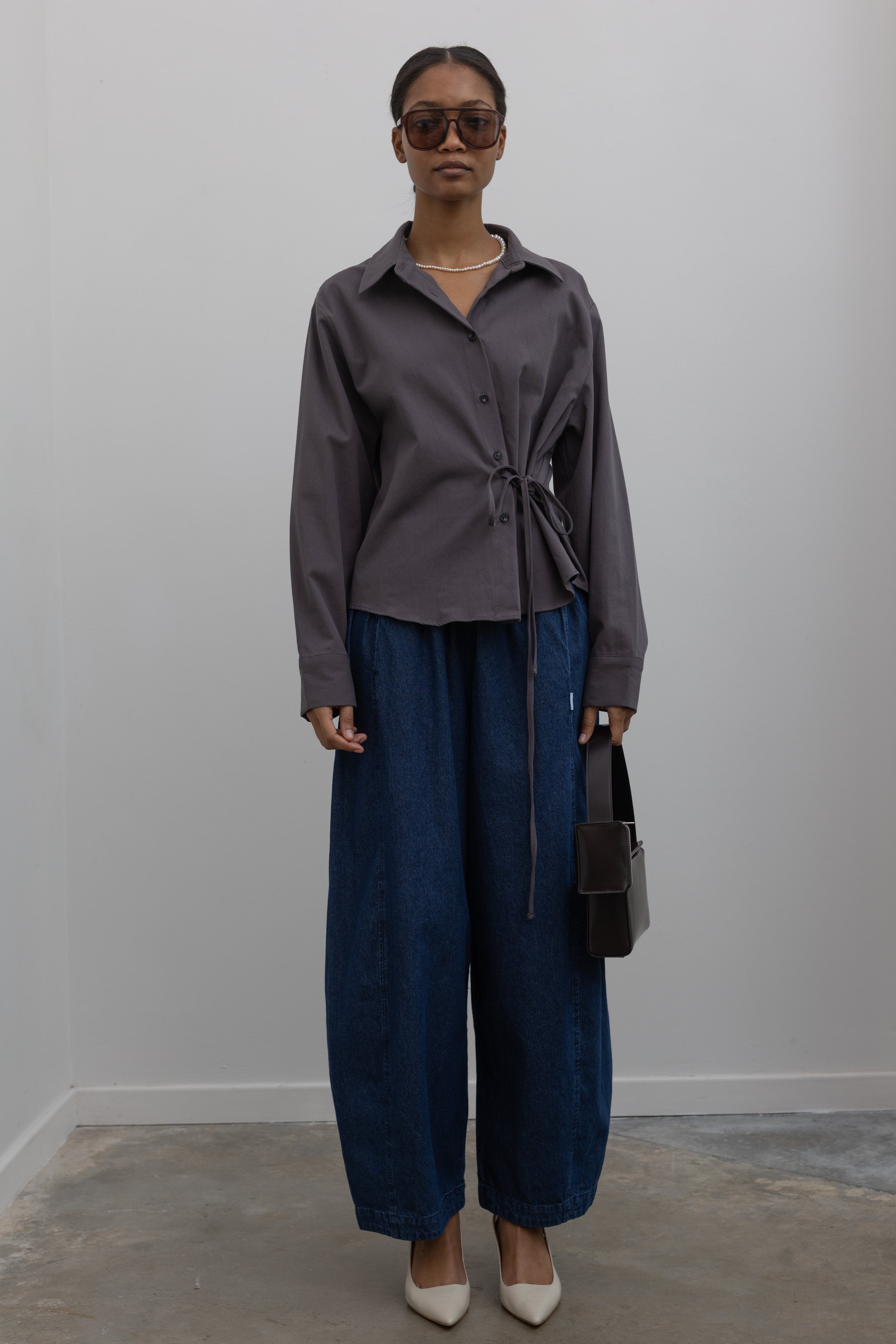 Woman wearing capsule wardrobe essential outfit in an Alas Eius label side tie button down shirt with Le Bon Shoppe Denim Blue Cotton Parachute Pants paired white leather slingback heels and a St. Agni shiny brown leather bag at Alas Eius. 