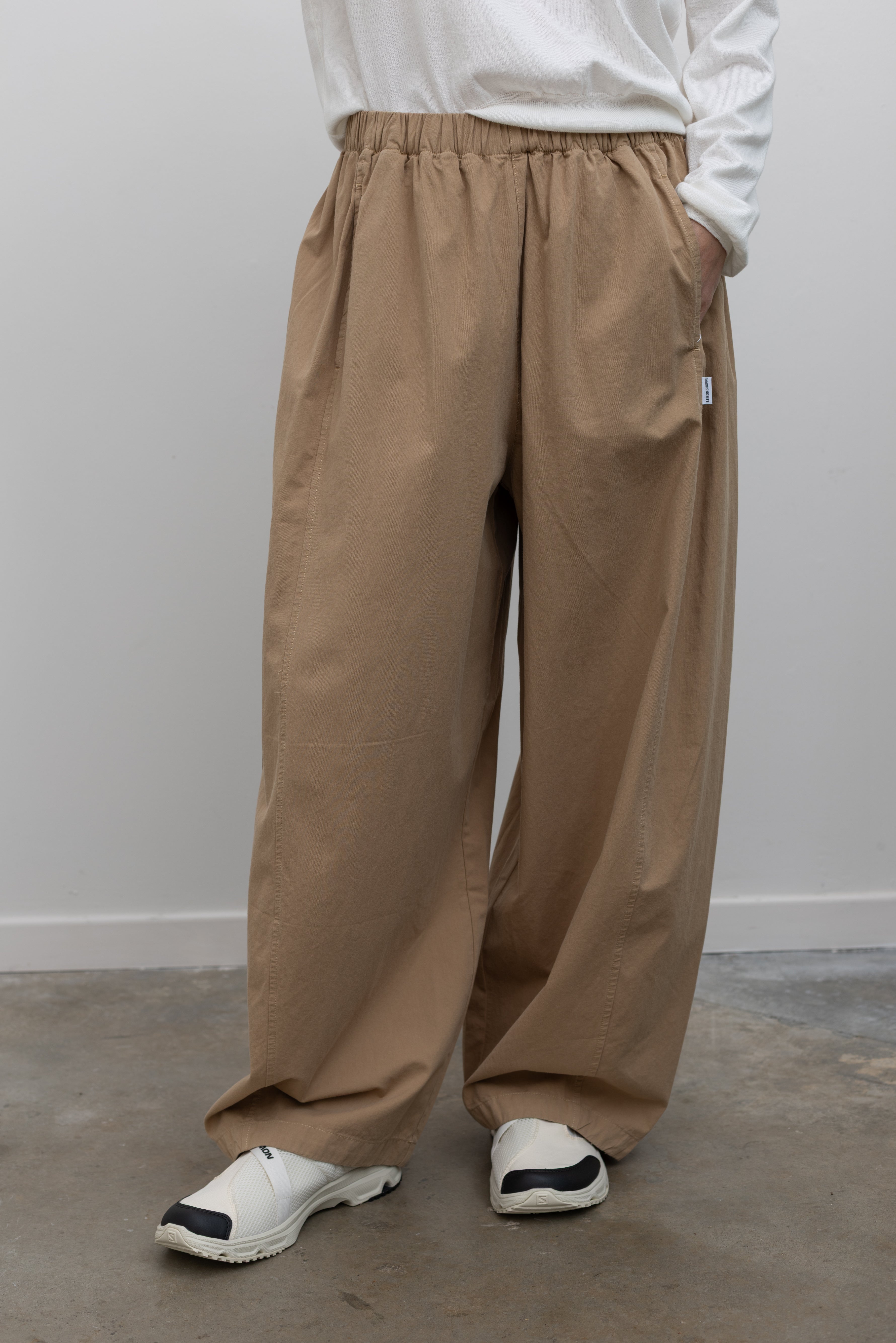Front view of a woman wearing elevated casual weekend fashion in the Le Bon Shoppe Khaki Cotton Parachute Pants featuring a lightweight cotton fabric, elastic waistband, and a voluminous oversized leg shape paired with Salomon sneakers and a white top at an Alas Eius store.