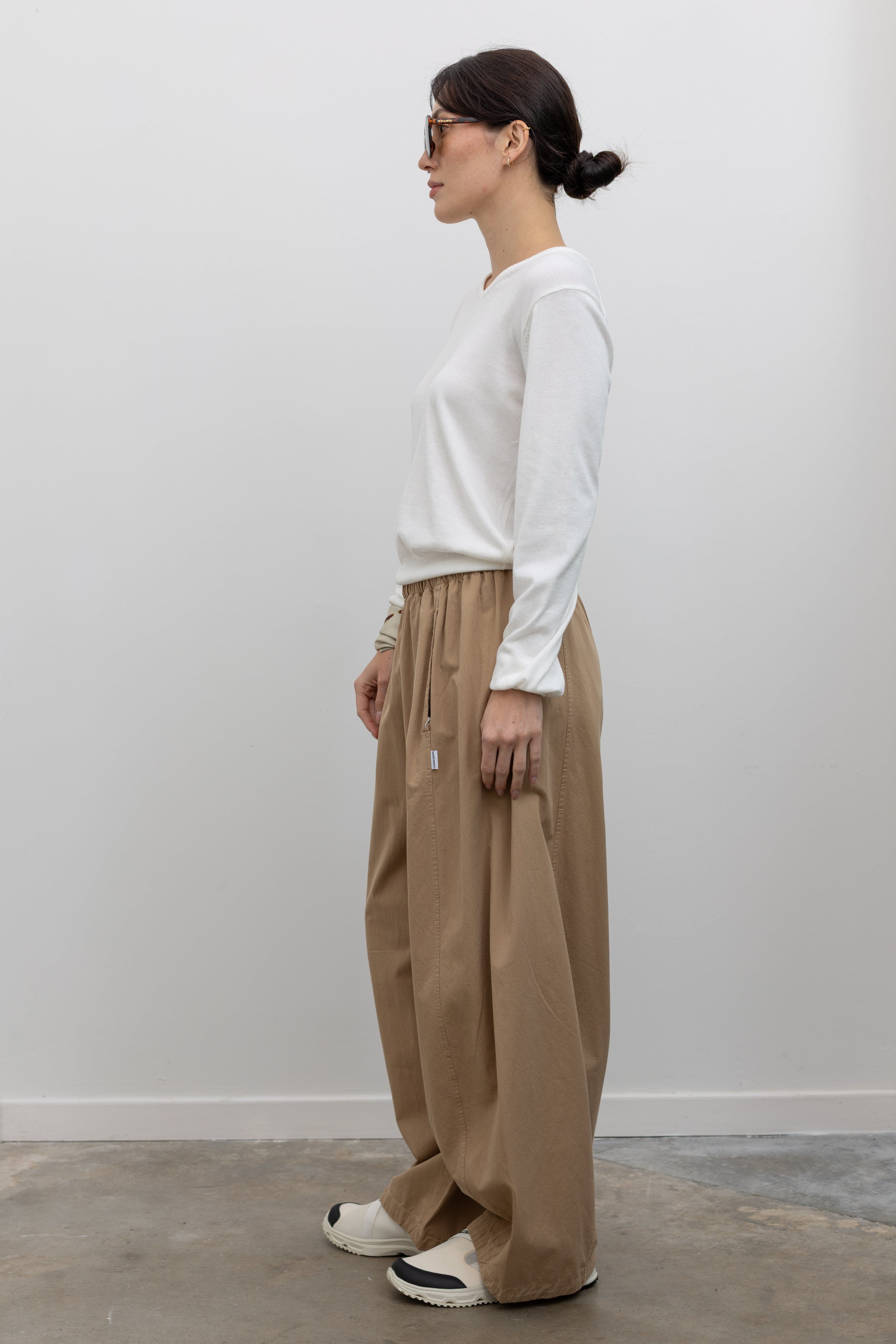 Full length front view of a woman wearing an elevated classic in the Le Bon Shoppe Khaki Cotton Parachute Pants featuring a lightweight cotton fabric, elastic waistband, and a voluminous oversized leg shape paired with Salomon sneakers at an Alas Eius store.