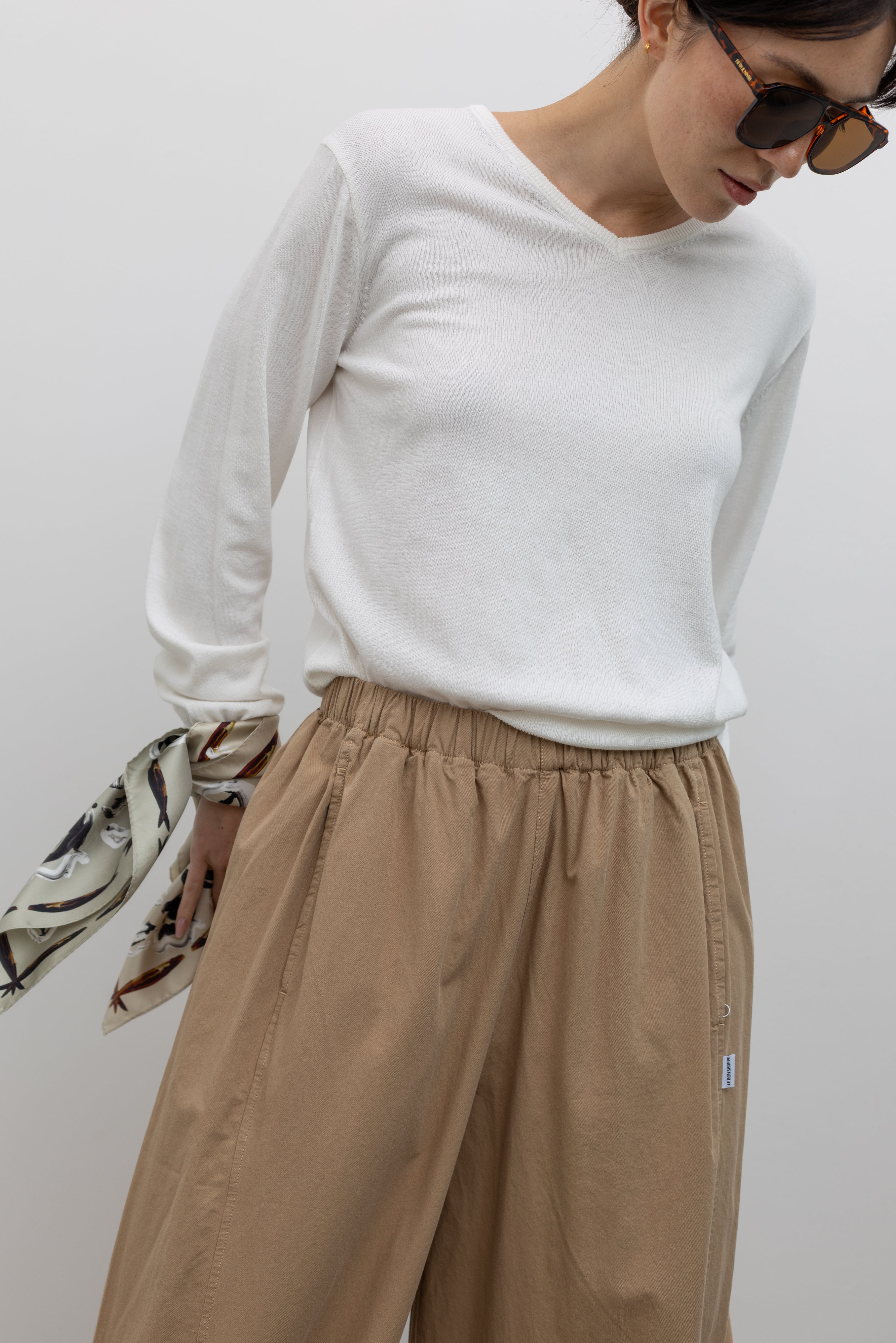 Front close-up view of a woman wearing elevated casual weekend fashion in the Le Bon Shoppe Khaki Cotton Parachute Pants featuring a lightweight cotton fabric, elastic waistband, and a voluminous oversized leg shape paired with Salomon sneakers and a white top at an Alas Eius store.