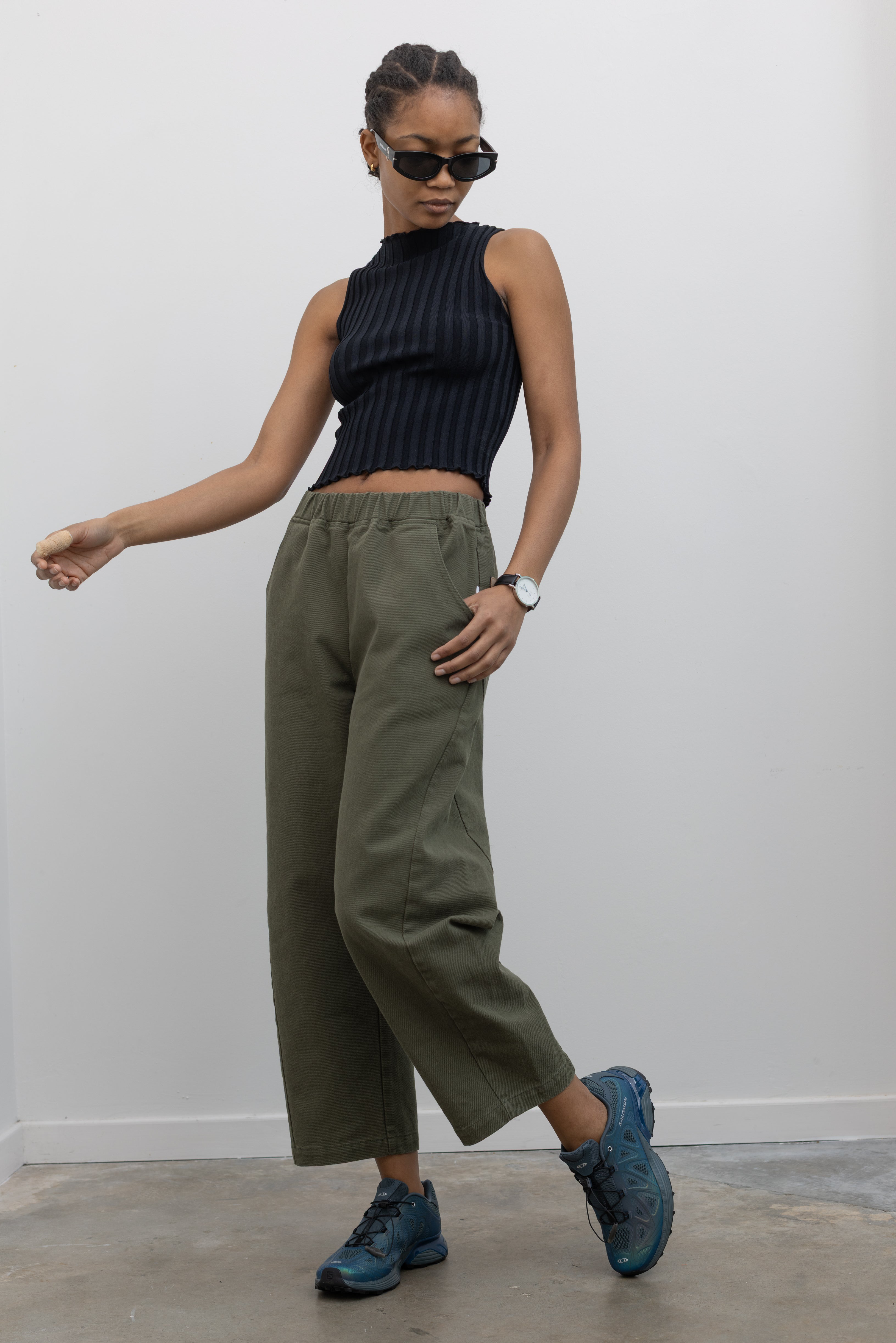 Woman wearing spring fashion in a pair of Le Bon Shoppe olive green curved arc pants with deep side seam pockets and an elastic waistband pair with a sleeveless ribbed knit top and Salomon sneakers at Alas Eius. 
