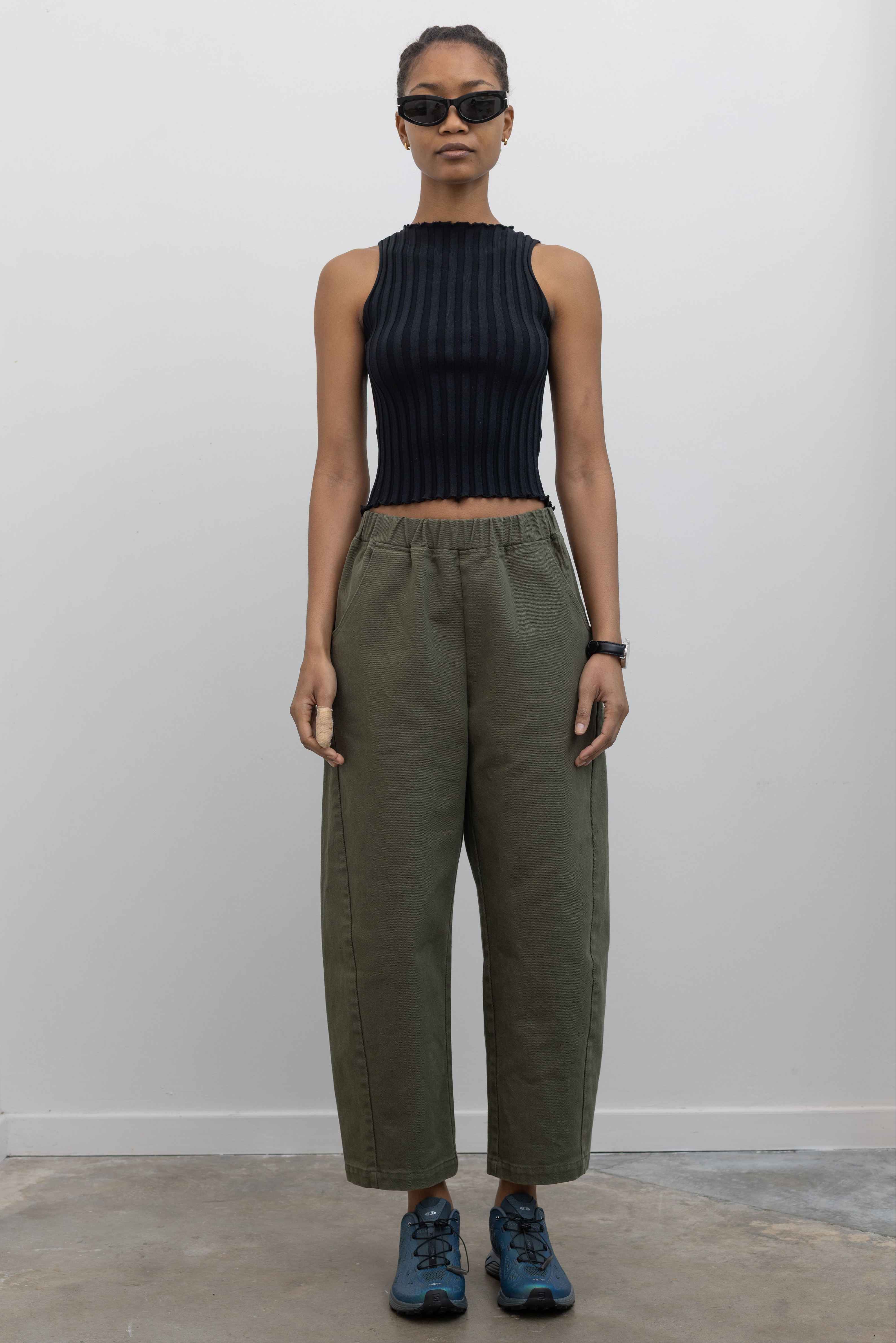 Full front view of a woman wearing spring fashion in a pair of Le Bon Shoppe olive green curved arc pants with deep side seam pockets and an elastic waistband pair with a sleeveless ribbed knit top and Salomon sneakers at Alas Eius. 