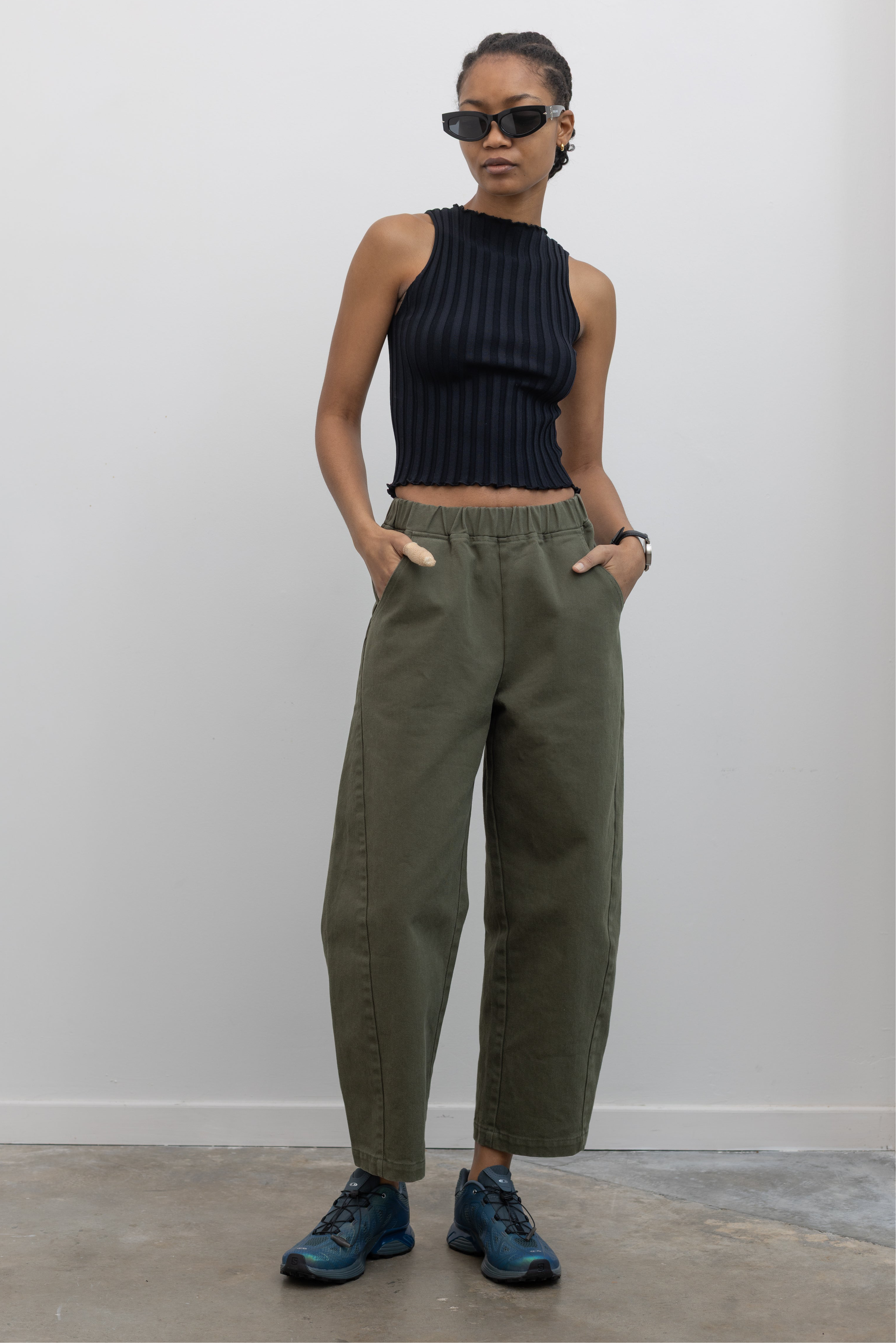 Woman wearing spring fashion in a pair of Le Bon Shoppe olive green curved arc pants with deep side seam pockets and an elastic waistband pair with a sleeveless ribbed knit top and Salomon sneakers at Alas Eius. 
