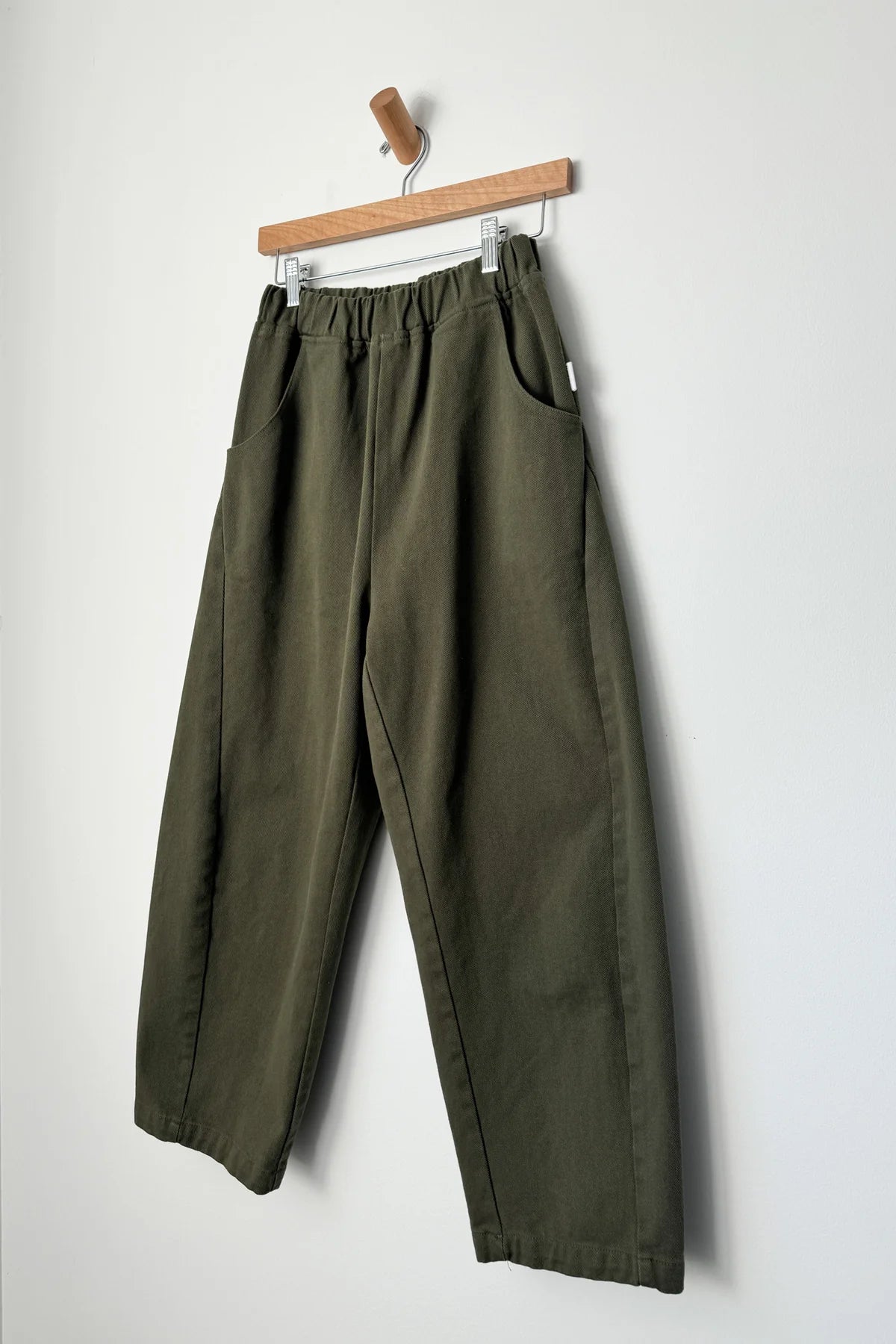 Casual spring fashion in a pair of Le Bon Shoppe olive green curved arc pants with deep side seam pockets and an elastic waistband on a wooden hanger at Alas Eius. 