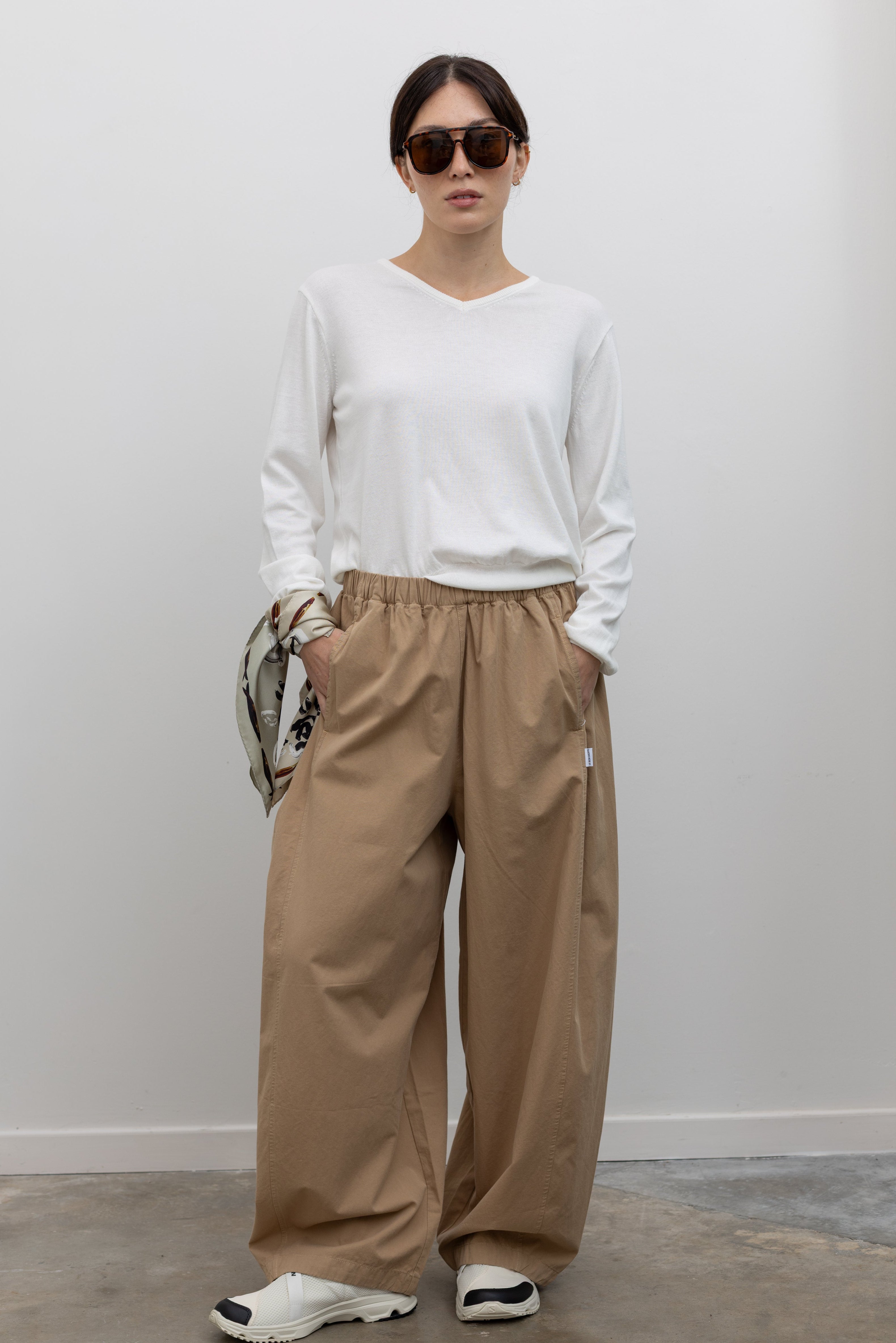 Full length front view of a woman wearing elevated casual weekend fashion in the Le Bon Shoppe Khaki Cotton Parachute Pants featuring a lightweight cotton fabric, elastic waistband, and a voluminous oversized leg shape paired with Salomon sneakers and a white top at an Alas Eius store.