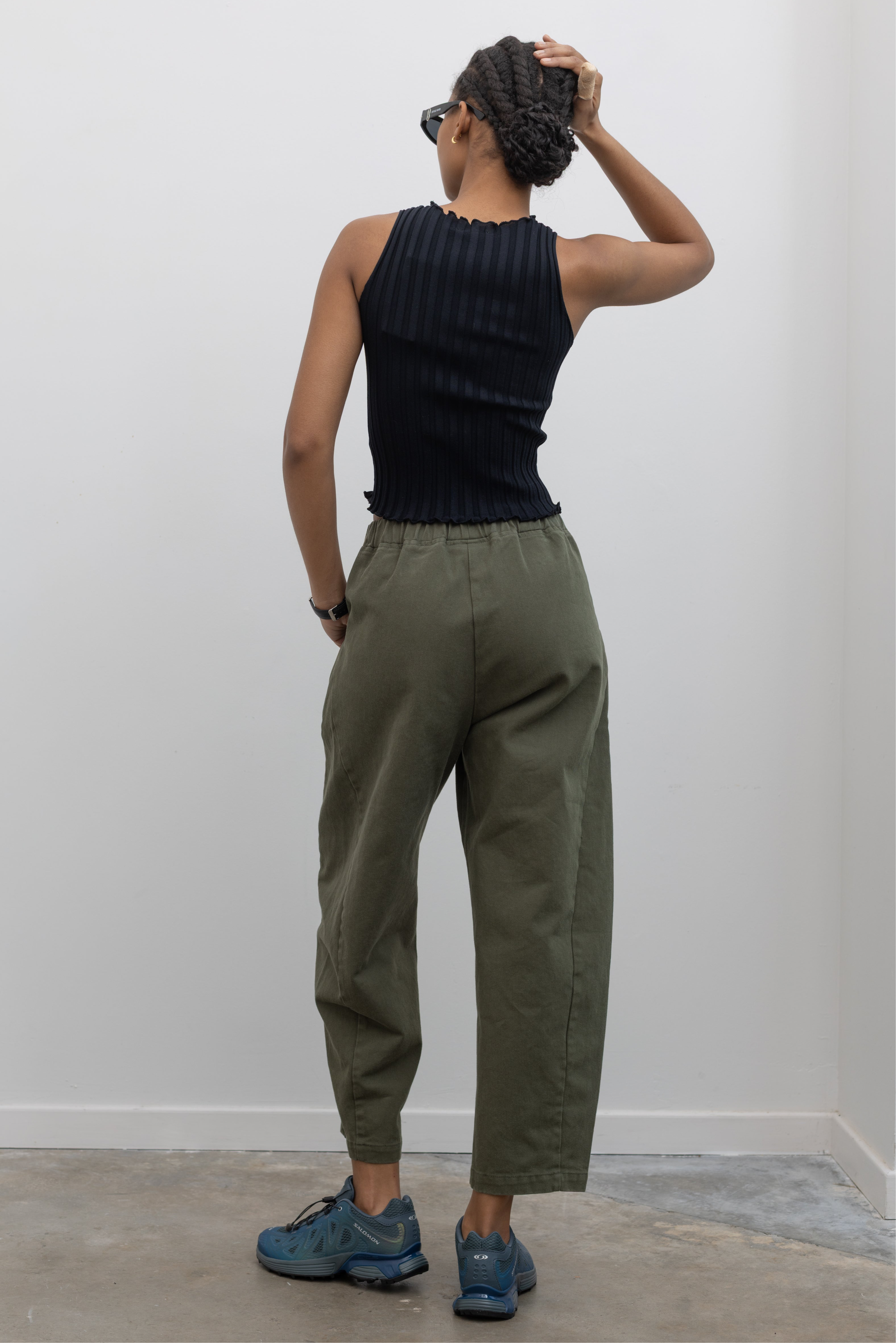 Back view of a woman wearing spring fashion in a pair of Le Bon Shoppe olive green curved arc pants with deep side seam pockets and an elastic waistband pair with a sleeveless ribbed knit top and Salomon sneakers at Alas Eius. 