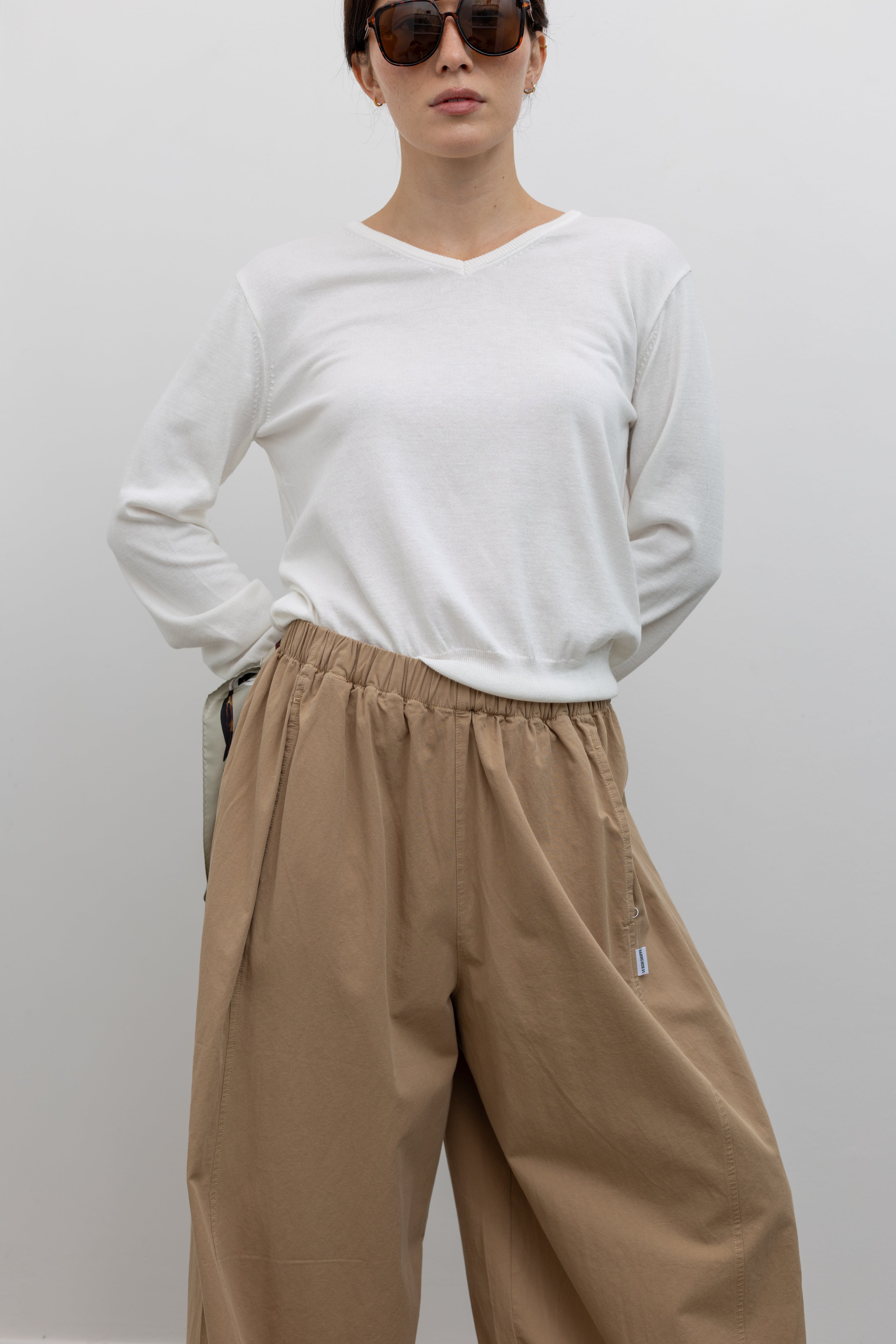 Front view of a woman wearing elevated casual weekend fashion in the Le Bon Shoppe Khaki Cotton Parachute Pants featuring a lightweight cotton fabric, elastic waistband, and a voluminous oversized leg shape paired with Salomon sneakers and a white top at an Alas Eius store.