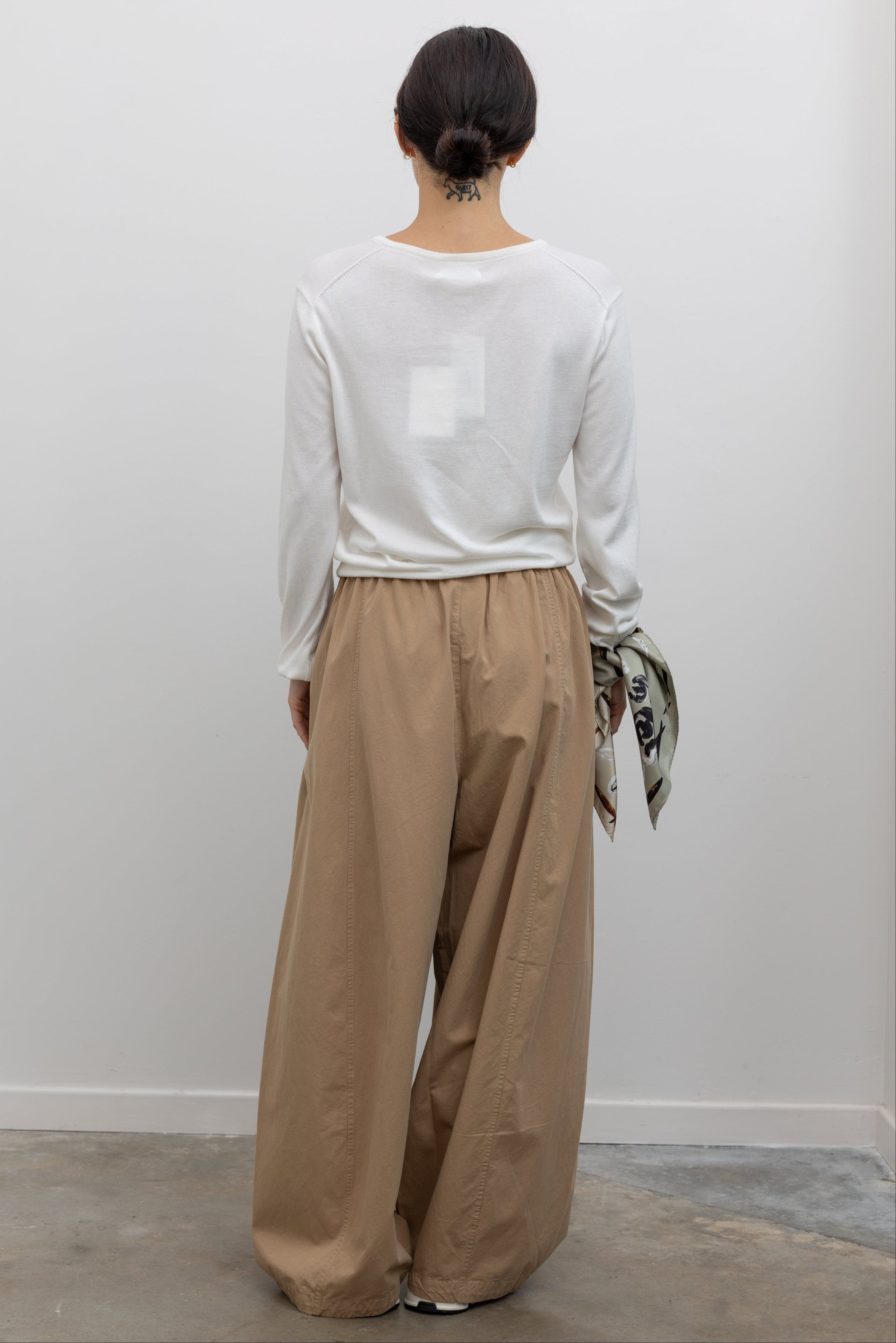 Full length back view of a woman wearing modern minimalist fashion in the Le Bon Shoppe Khaki Cotton Parachute Pants featuring a lightweight cotton fabric, elastic waistband, and a voluminous oversized leg shape paired with Salomon sneakers and a white top at an Alas Eius store.