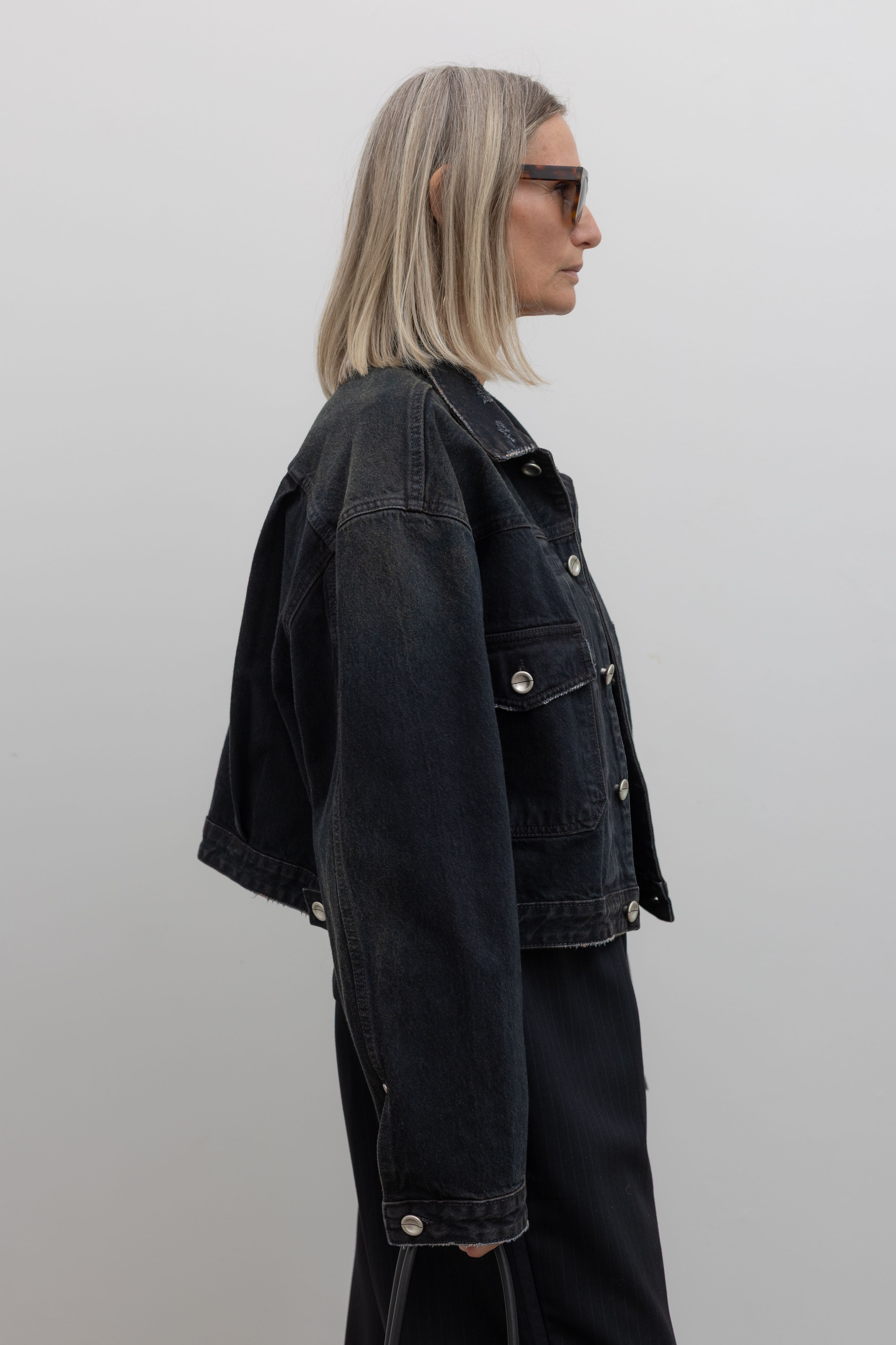 Side view of a woman wearing Low Classic Mahogany Black Denim Short Trucker Jacket featuring silver hardware and a relaxed cropped fit at Alas Eius. 