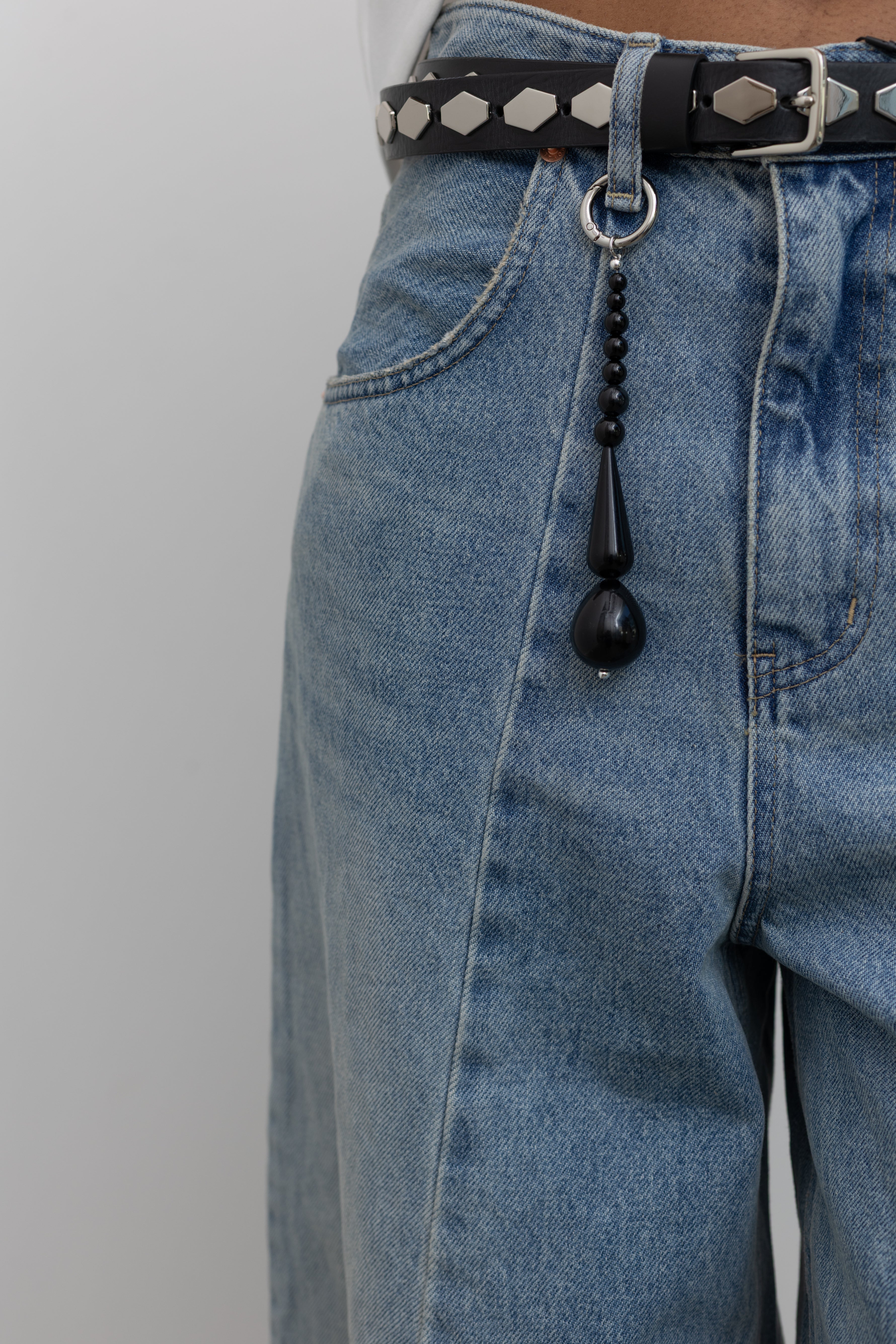 Close-up view of modern Maslo black bead bag charm hooked on a belt loop of light wash denim jeans and paired with a silver studded black leather belt at Alas Eius. 