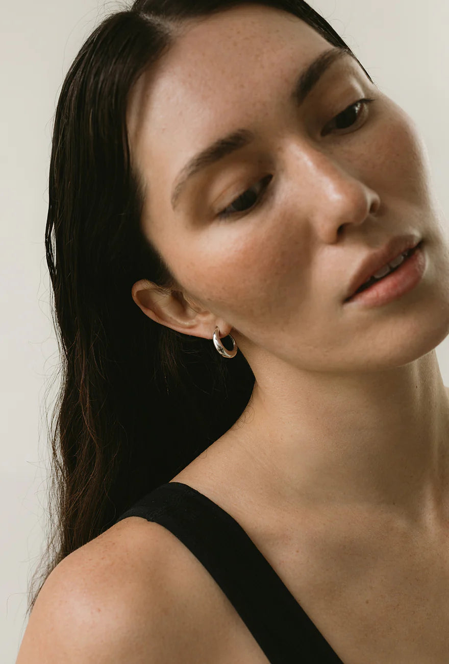 Woman wearing minimalist jewelry in a pair of Maslo Ceres sterling silver thick hoop stud earrings at Alas Eius. 