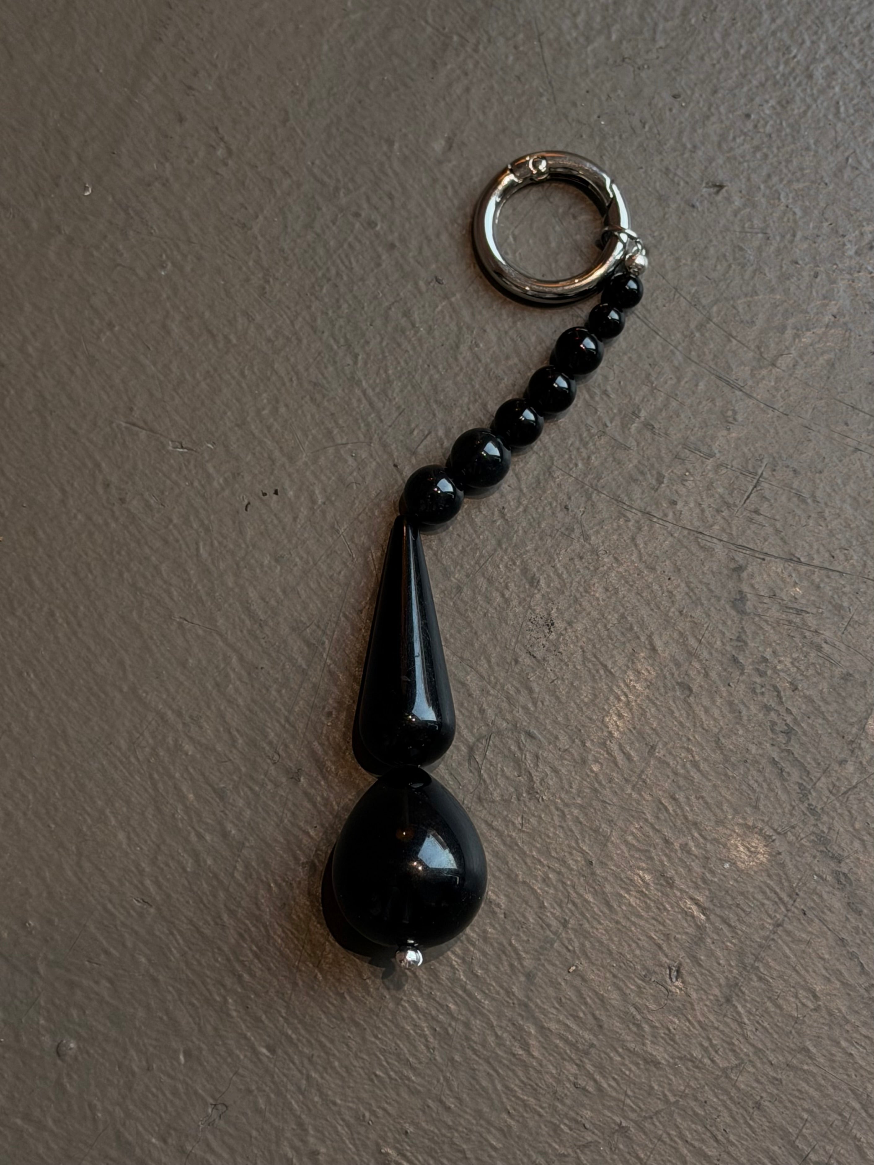 Close-up view of modern Maslo black bead bag charm hooked on a belt loop of light wash denim jeans and paired with a silver studded black leather belt at Alas Eius. 