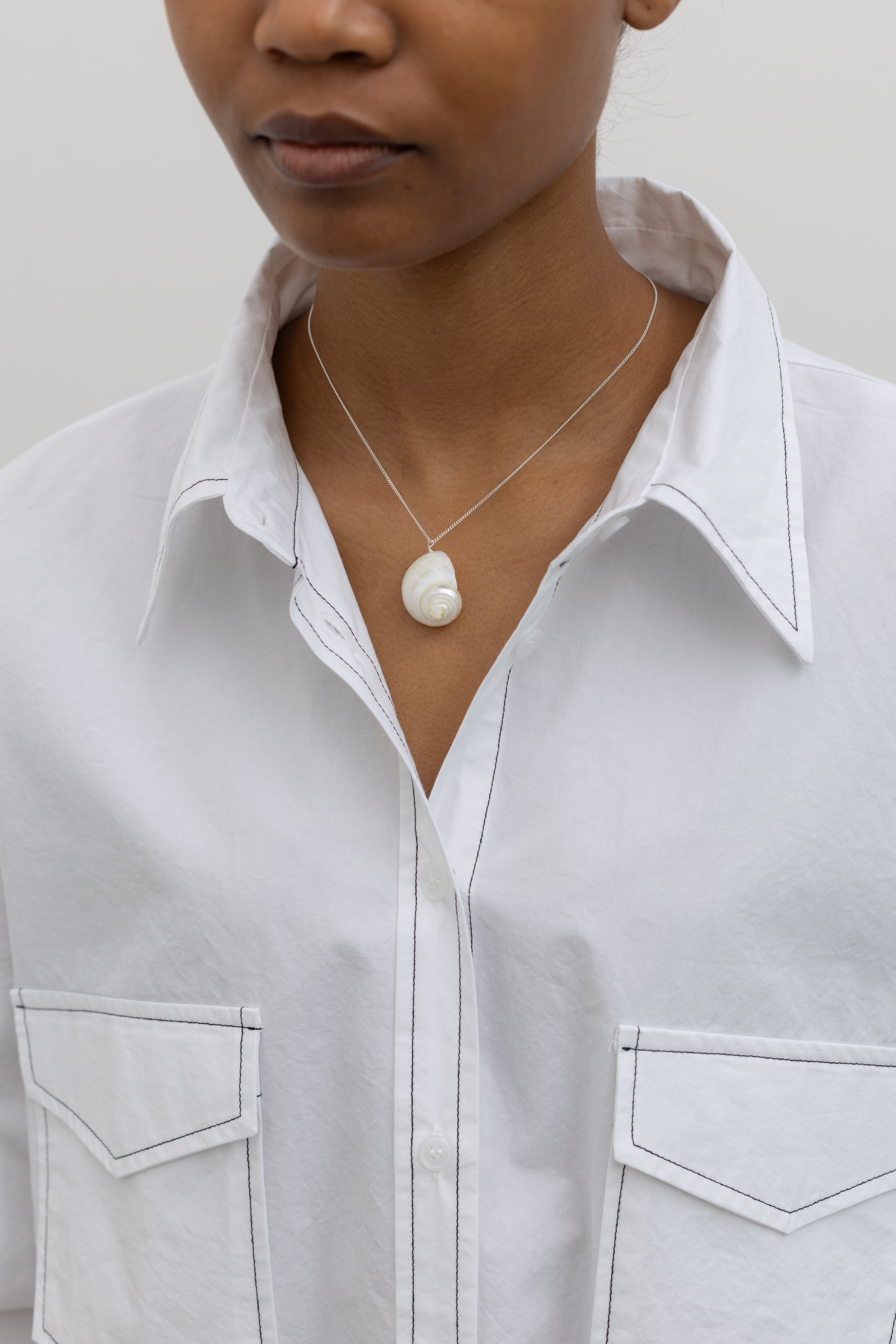 Woman wearing a Maslo necklace featuring a white shell charm pendant on a fine sterling silver chain paired with an Alas Eius label white button down shirt with black contrast stitching. 
