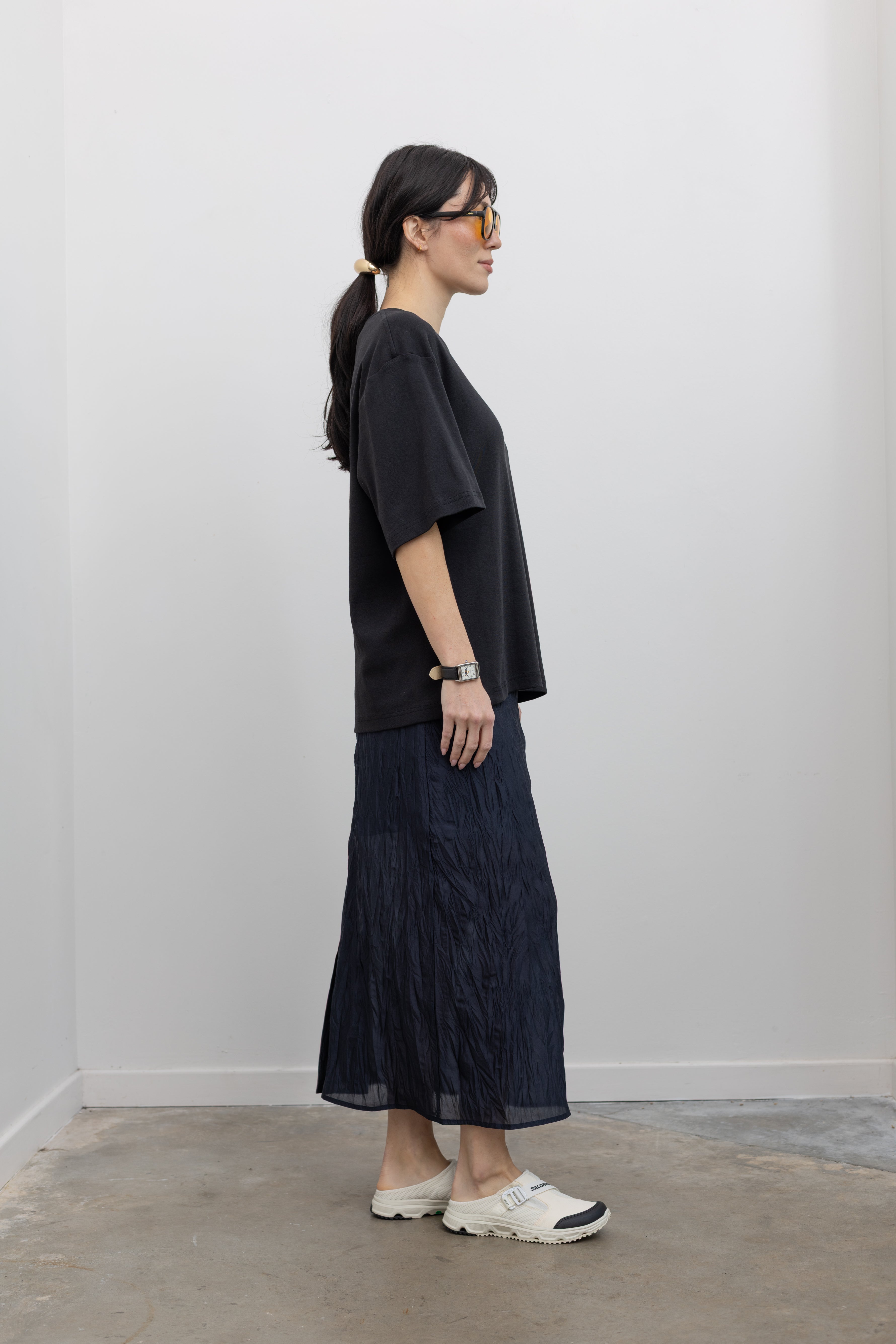 Full length side view of a woman standing wearing contemporary spring fashion in a Mijeong Park navy blue lightweight, semi-sheer crinkled fabric lined midi skirt paired with a Mijeong Park black oversized t-shirt and Salomon sneakers at an Alas Eius store. 