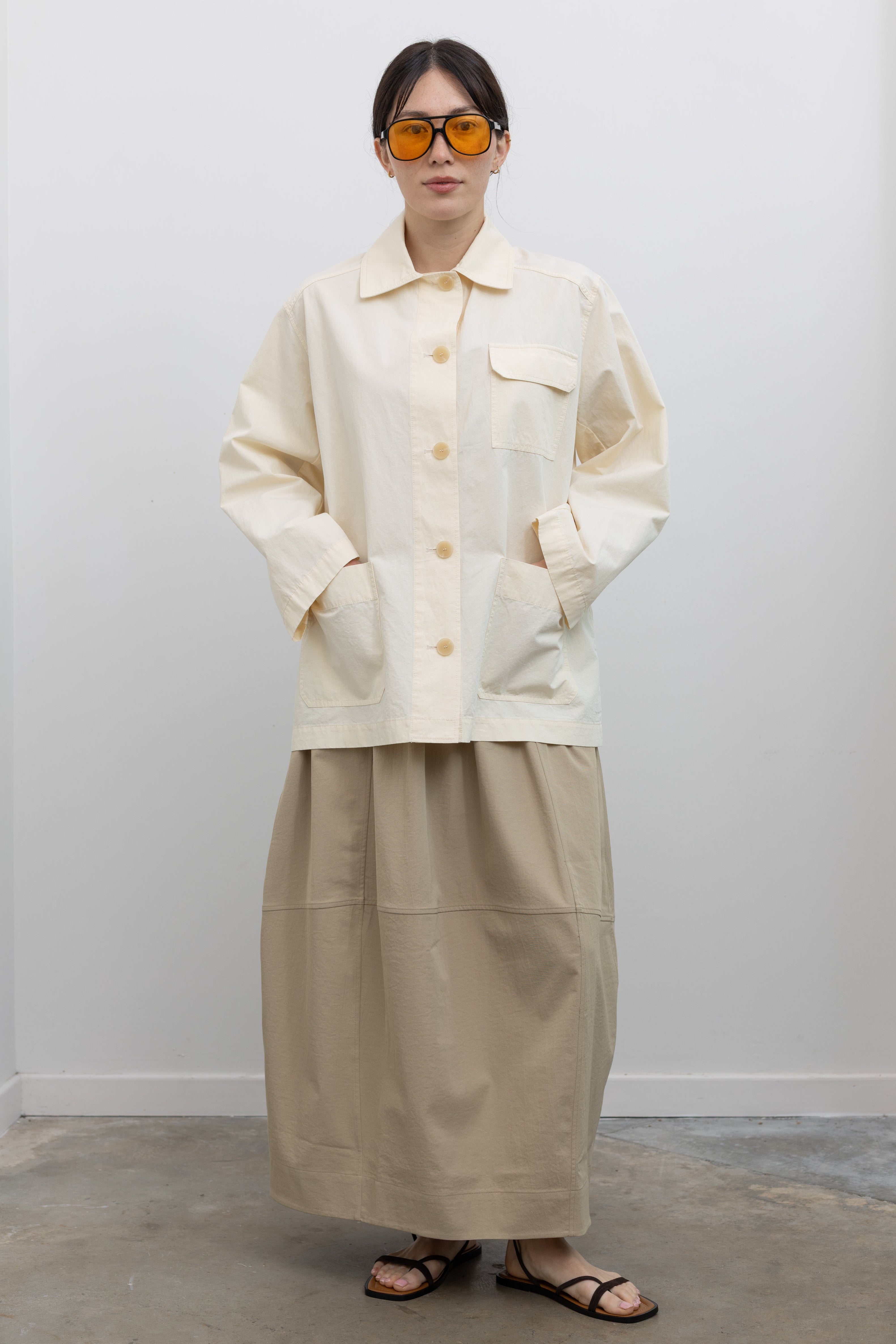 Full length front view of a woman standing with hands in front pockets wearing chic and modest Spring fashion in a Mijeong Park beige balloon midi skirt paired with a cream colored linen blend work jacket featuring a relaxed fit, two front patch pockets and a button down front closure for versatile styling at an Alas Eius store. 