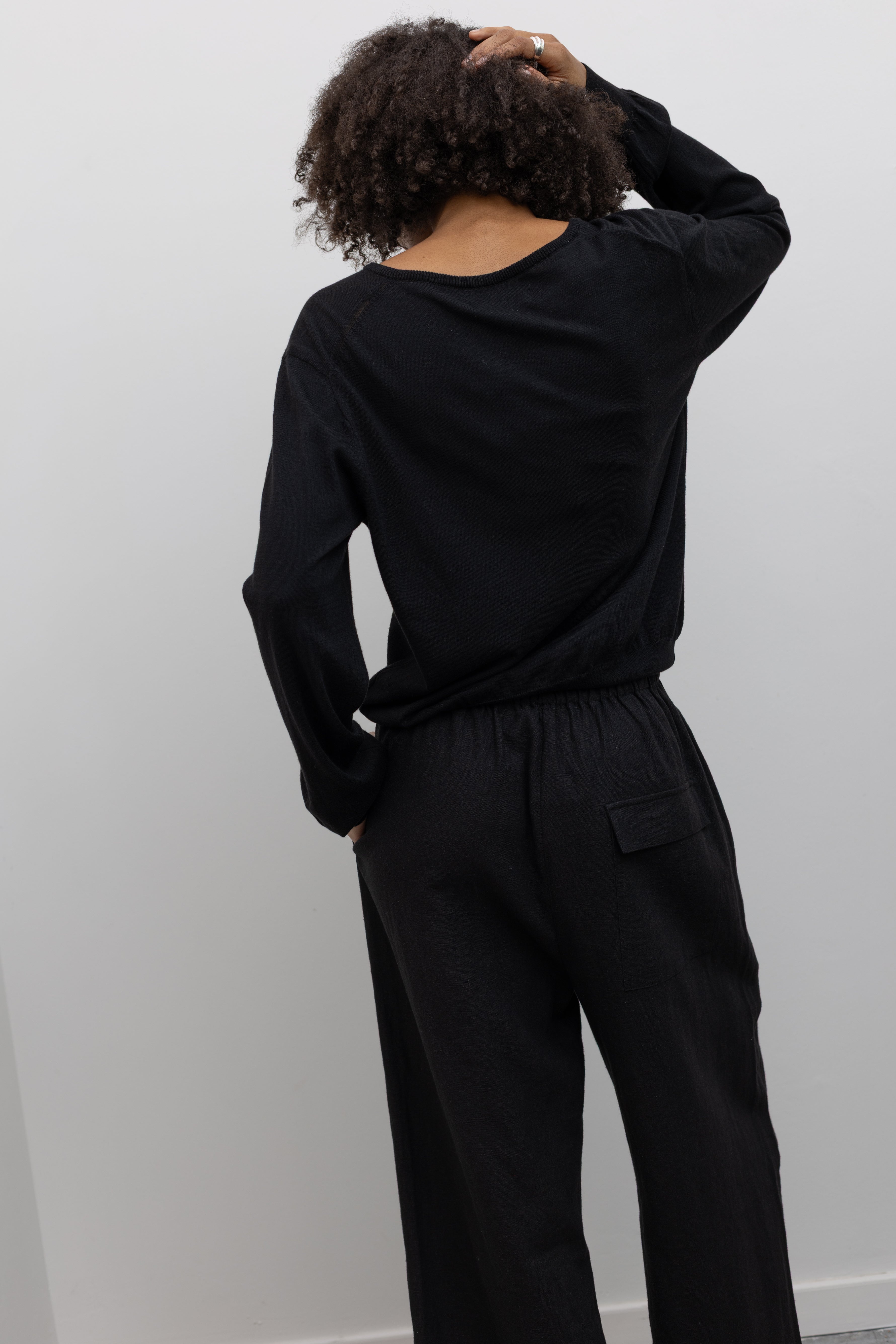 Back view of a woman standing wearing elevated casual weekend fashion in a Mijeong park Black Cotton Blend V-Neck Long Sleeve Knit Top paired with Mijeong Park Black Linen Blend Cropped Pants featuring a comfortable drawstring waist, front and back patch pockets, and a cropped length suitable for a minimalist, versatile wardrobe at an Alas Eius store.