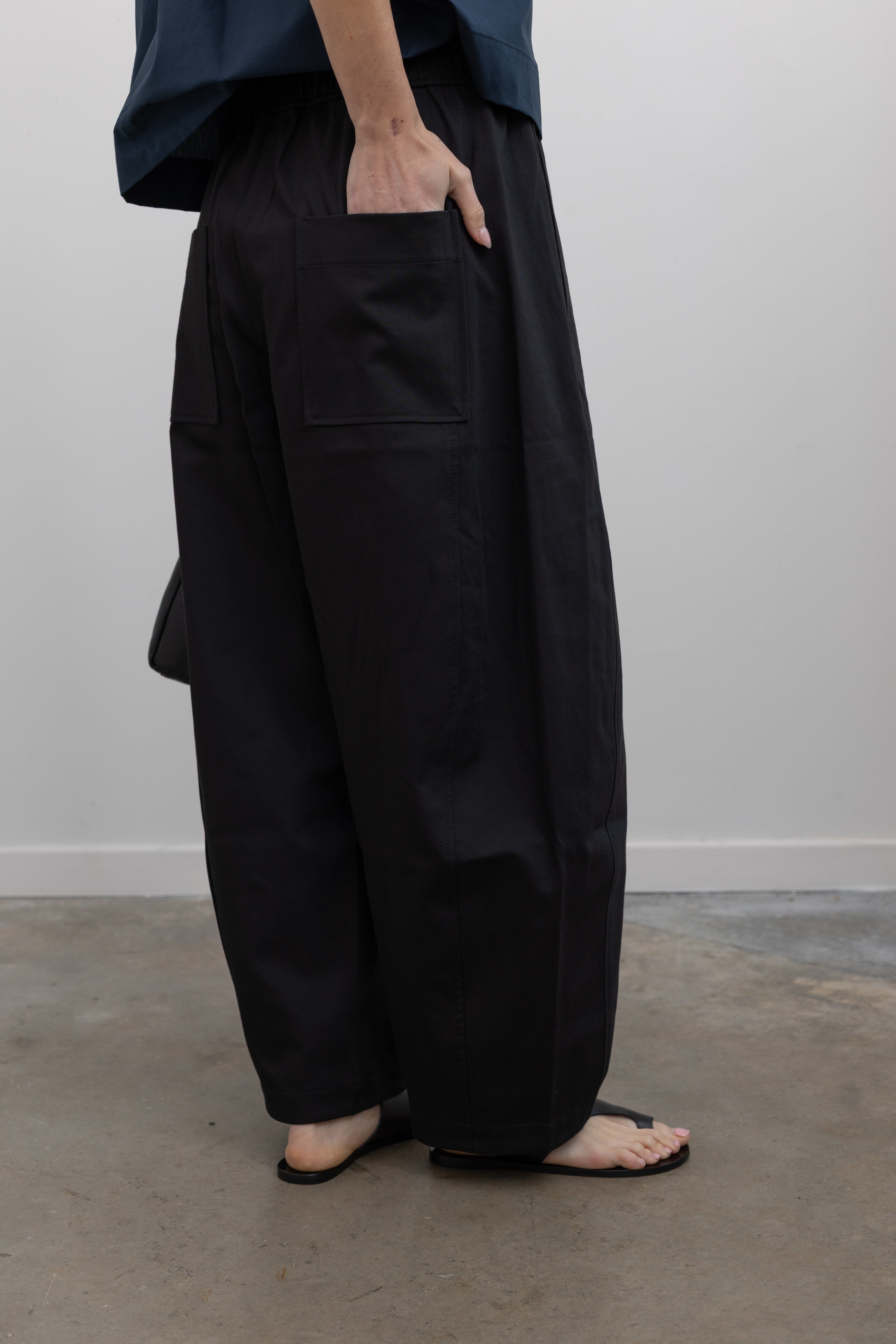Close-up back view of contemporary womenswear spring fashion in Mijeong Park black baggy balloon pants with curved outseams, a relaxed fit, elastic waistband, and back patch pockets paired with a structured navy blue short sleeve cropped hem button down shirt at an Alas Eius store. 