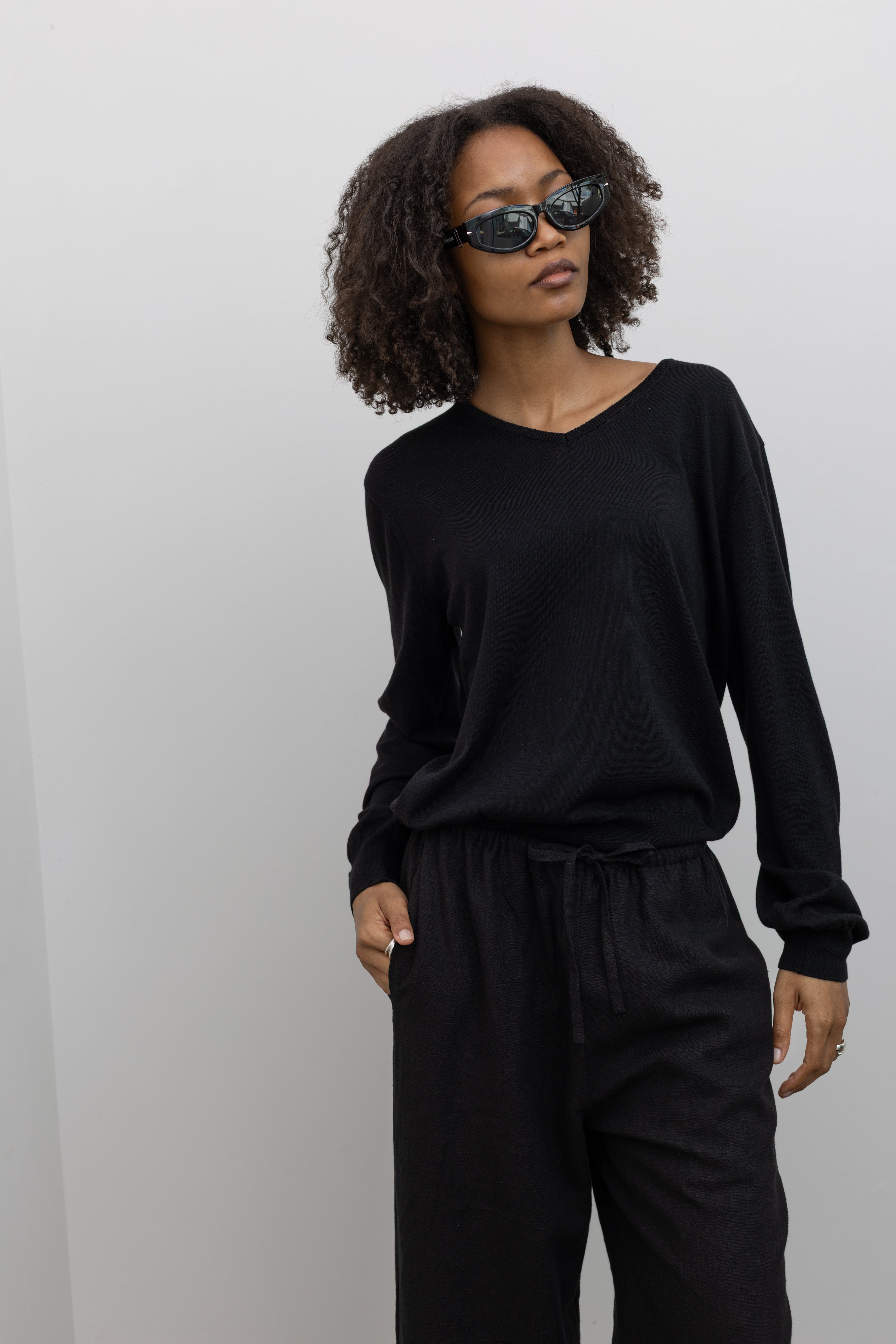 Front view of a woman standing wearing elevated casual weekend fashion in a Mijeong park Black Cotton Blend V-Neck Long Sleeve Knit Top paired with Mijeong Park Black Linen Blend Cropped Pants featuring a comfortable drawstring waist, front and back patch pockets, and a cropped length suitable for a minimalist, versatile wardrobe at an Alas Eius store.