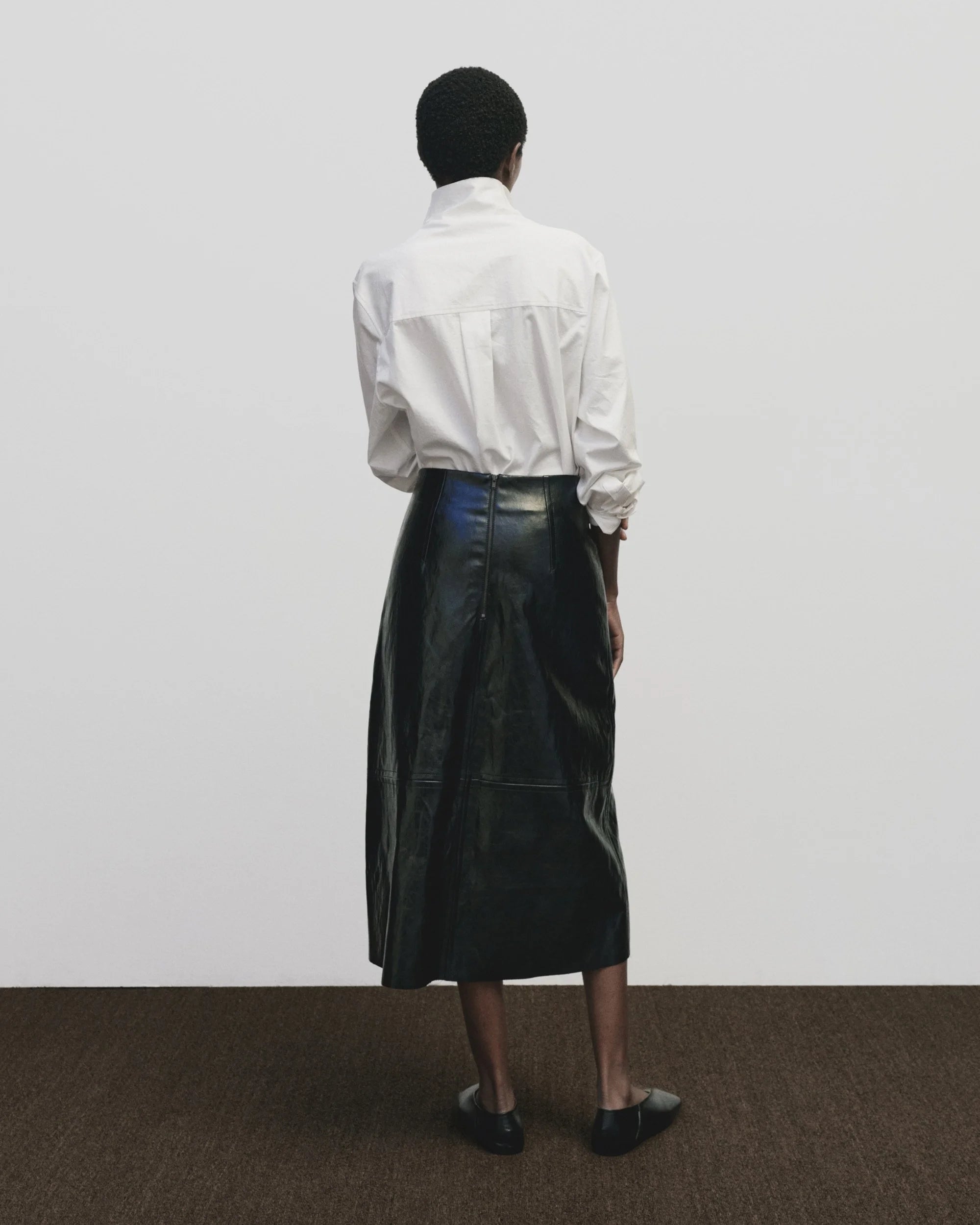 Person wearing a white shirt and black leather skirt standing against a plain white wall.
