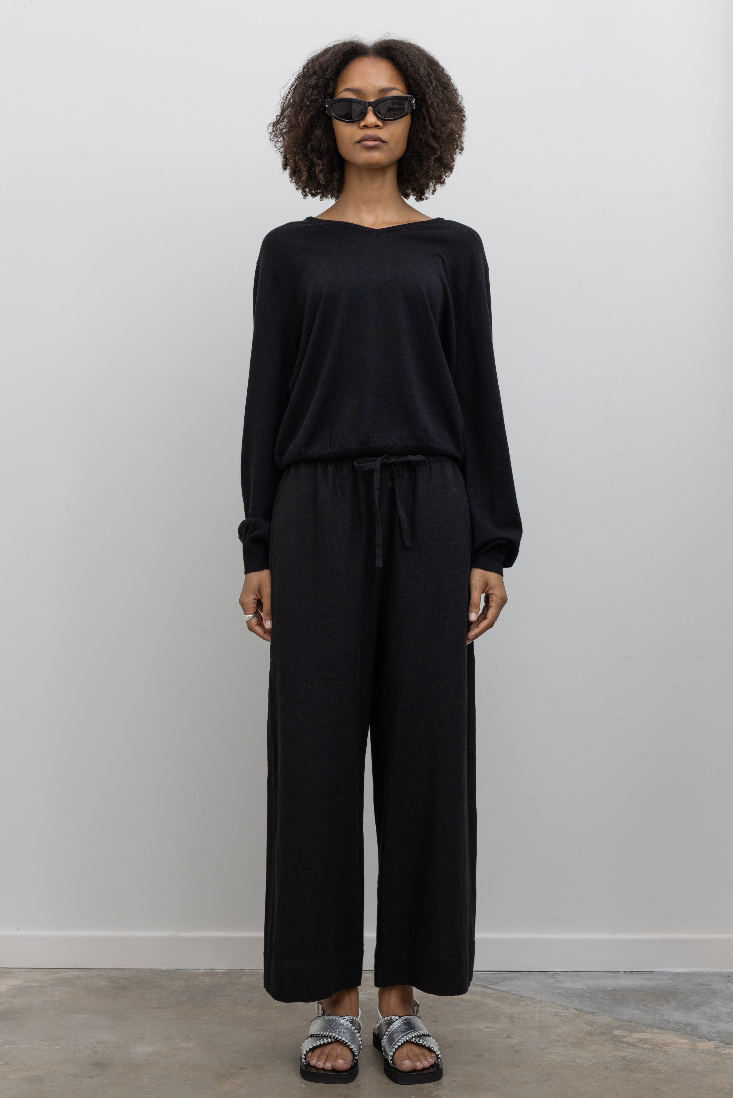 Full length front view of a woman standing wearing elevated casual weekend fashion in a Mijeong park Black Cotton Blend V-Neck Long Sleeve Knit Top paired with Mijeong Park Black Linen Blend Cropped Pants featuring a comfortable drawstring waist, front and back patch pockets, and a cropped length suitable for a minimalist, versatile wardrobe at an Alas Eius store.