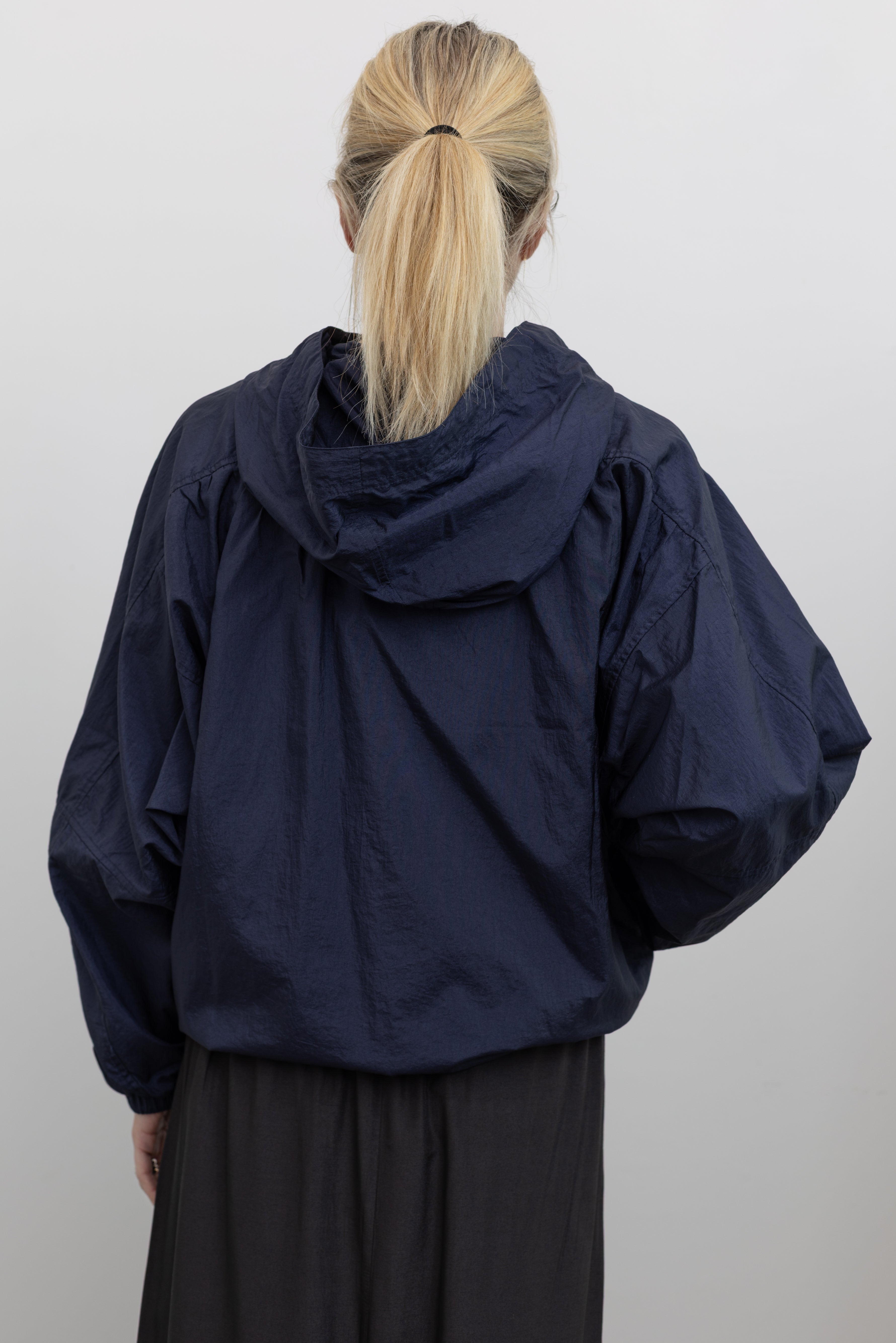 Close-up back view of a woman wearing an elevated outerwear staple in a  Mijeong Park navy blue nylon shirred anorak jacket featuring an oversized fit, front pockets, hood, and a zipper closure at an Alas Eius store. 