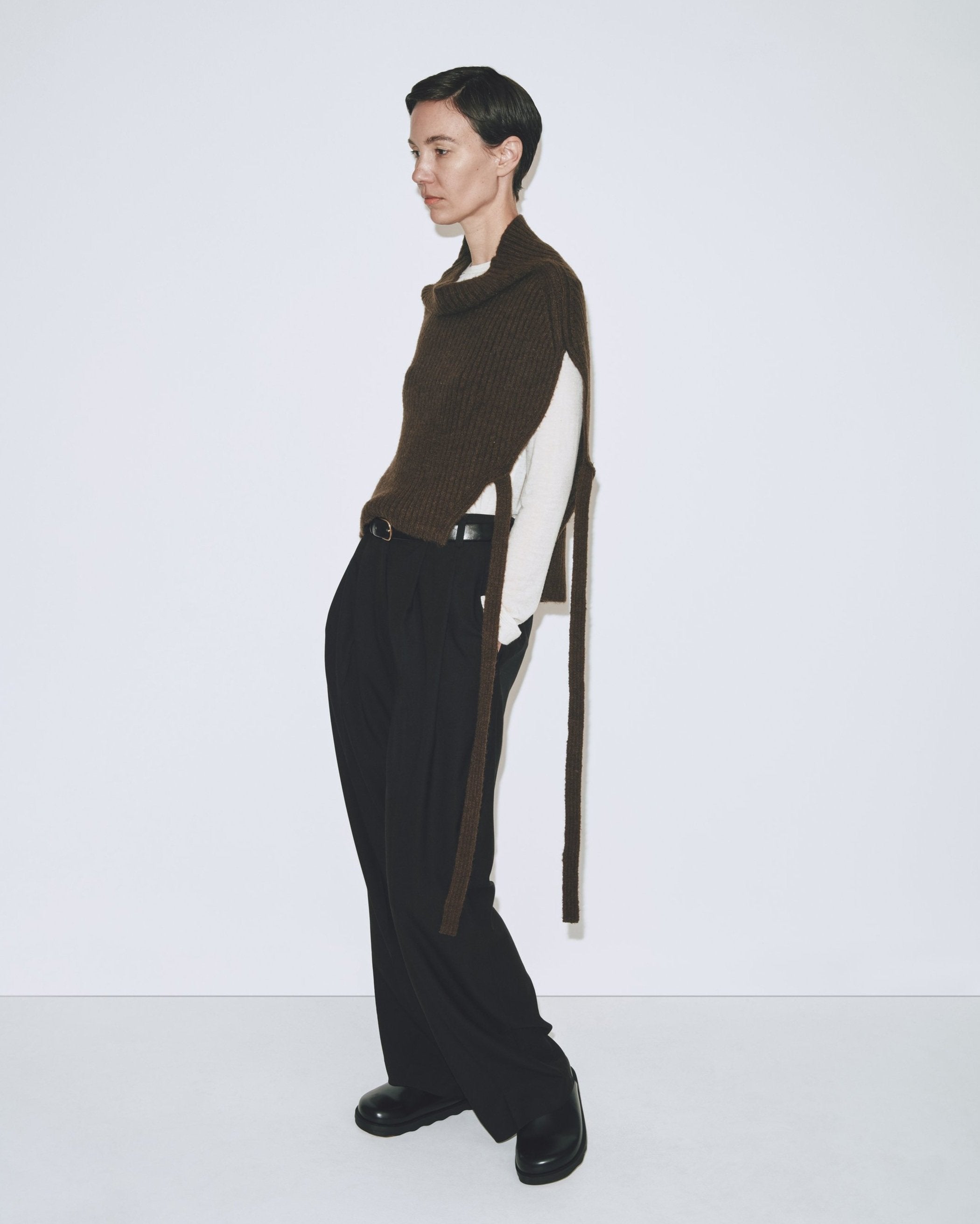 A person wearing an olive brown ribbed knitted Mijeong Park neck warmer with a mock neck neckline and tie waist detail, paired with a black pants at Alas Eius