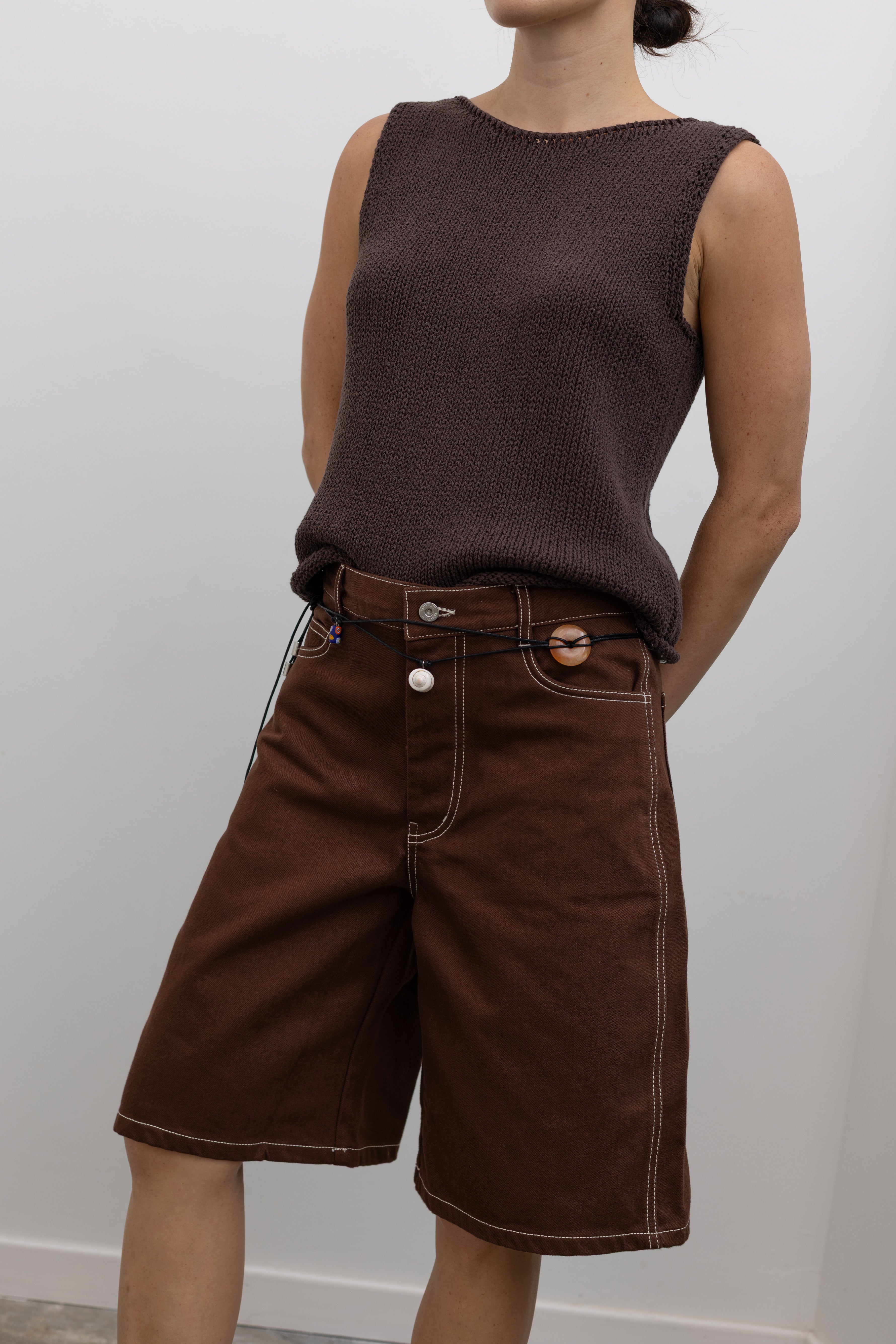 Close-up front view of a woman posing wearing modern minimalist spring and summer fashion in a Mijeong Park dark brown boat neck knit tank featuring a relaxed fit and rolled edges at the neck and hem paired with Kowtow Framework Organic Cotton Brown Denim Long Shorts that sit just above the knee and feature white contrast stitching at an Alas Eius store.  