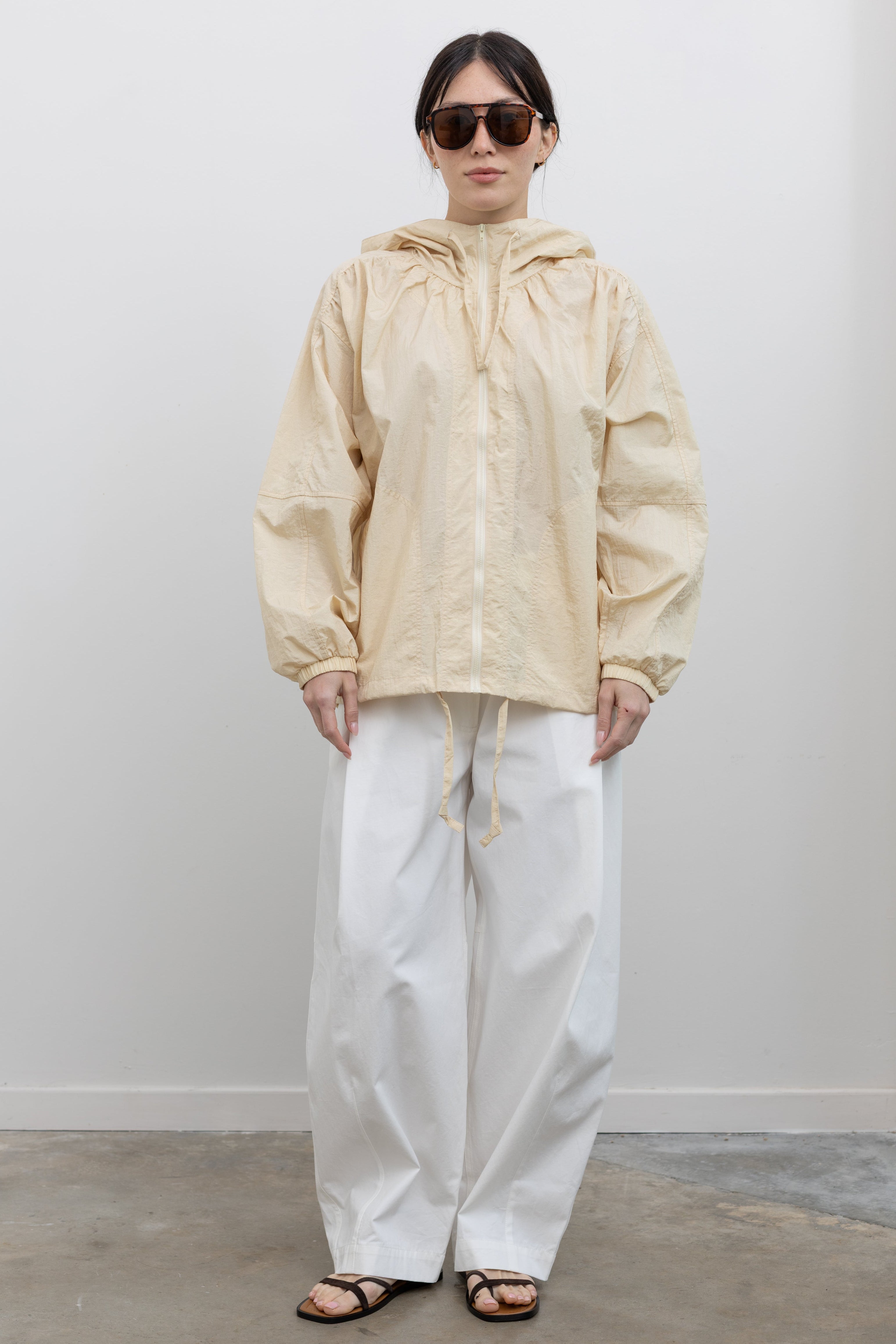 Full length front view of a woman standing wearing spring fashion in Mijeong Park white 100% cotton barrel leg pants featuring curved outseams that create architectural volume and a partially elasticated waistband and belt loops for a tailored look paired with a Mijeong Park cream colored shirred anorak jacket at an Alas Eius store. 