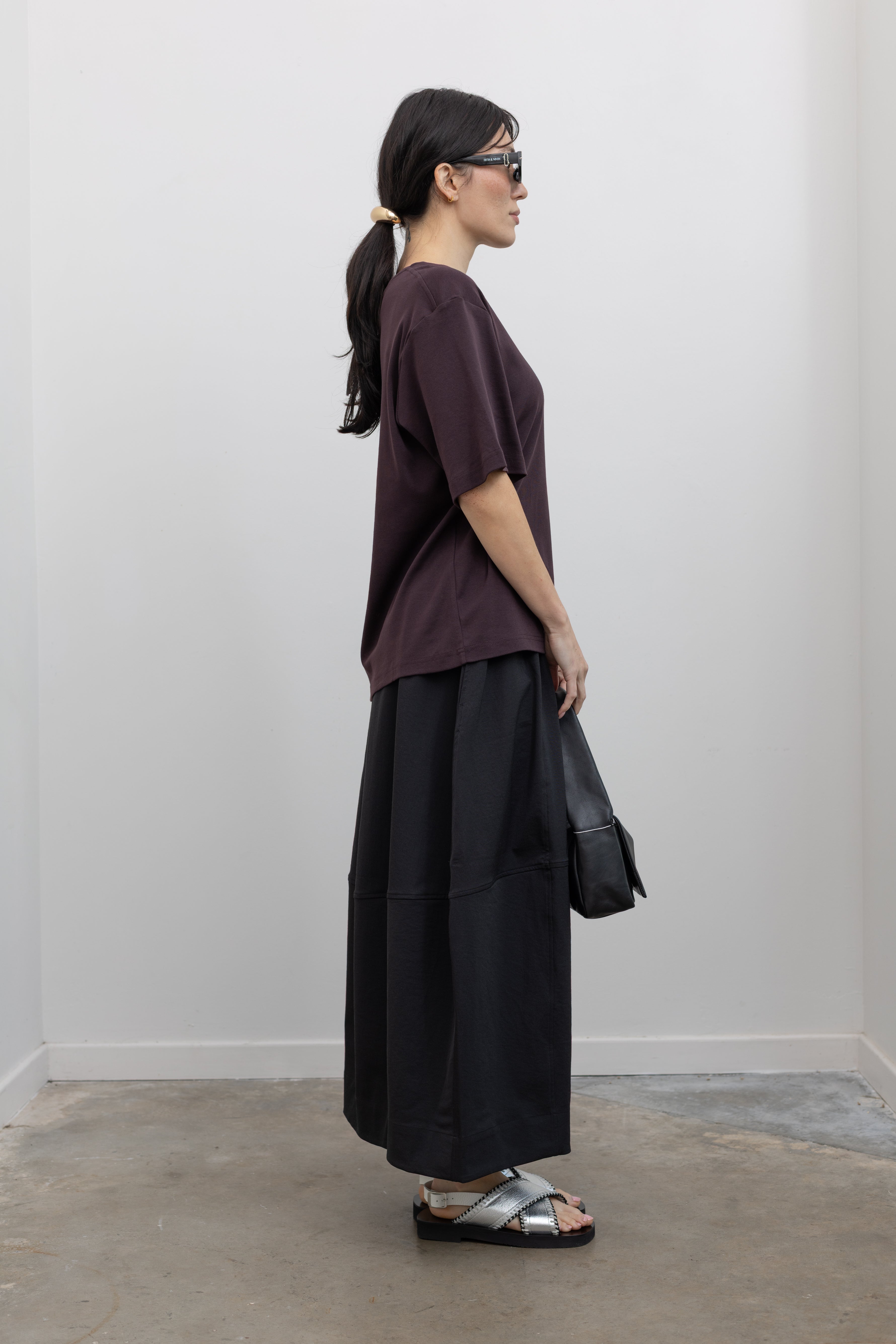Full length side view of a woman standing wearing contemporary womenswear spring fashion in a Mijeong Park dark brown oversized t-shirt with a black voluminous balloon midi-to-maxi silhouette skirt featuring an elastic waistband paired with silver metallic slip-on sandals and a sleek St. Agni black leather shoulder bag at an Alas Eius store. 