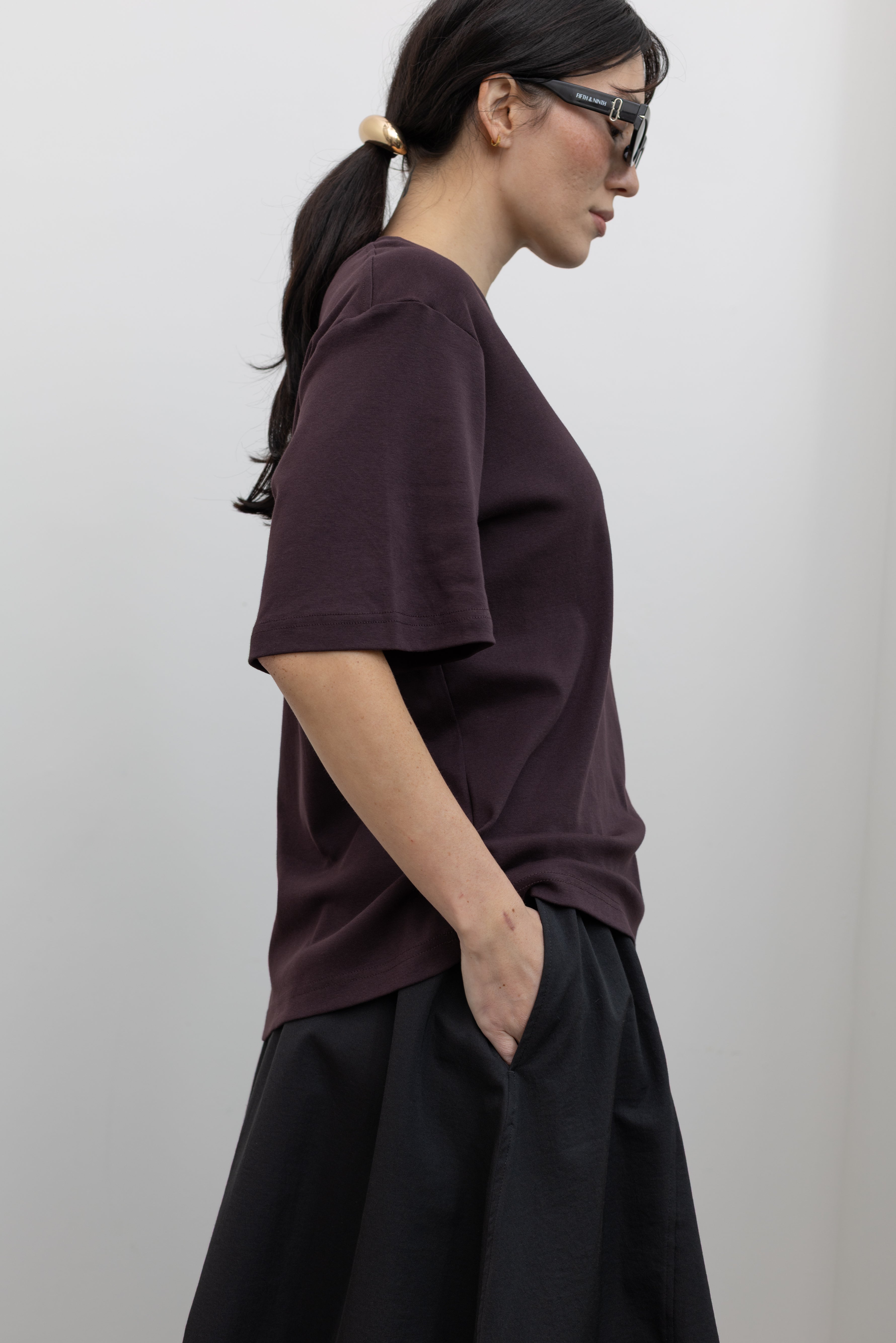 Side view of a woman standing wearing chic weekend and travel capsule staples in a Mijeong Park dark brown oversized t-shirt with a dropped shoulder paired with an Alas Eius label black nylon skirt and a modern leather St. Agni bag at an Alas Eius store. 