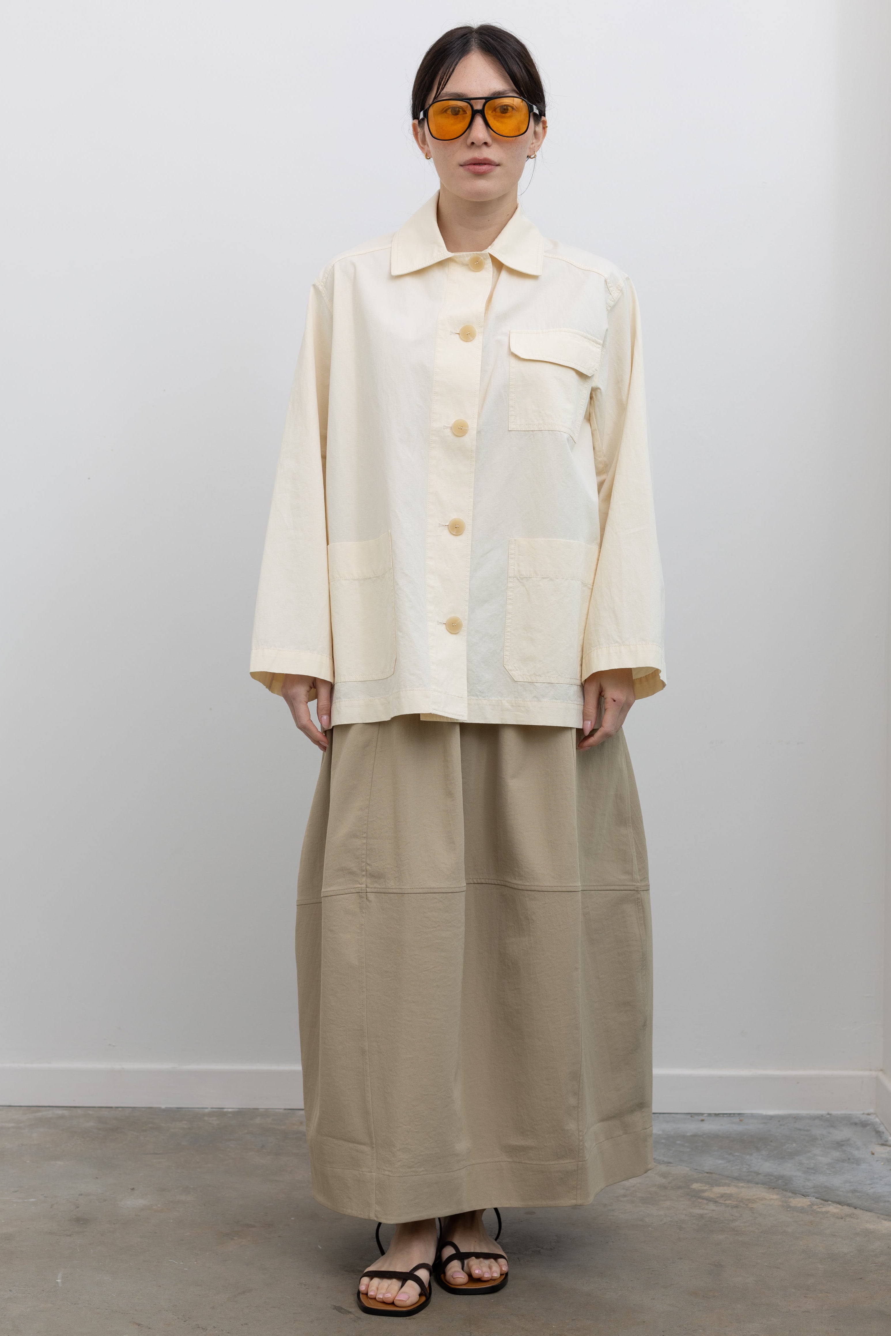 Full length front view of a woman standing wearing chic and modest Spring fashion in a Mijeong Park beige balloon midi skirt paired with a cream colored linen blend work jacket featuring a relaxed fit, two front patch pockets and a button down front closure for versatile styling at an Alas Eius store. 