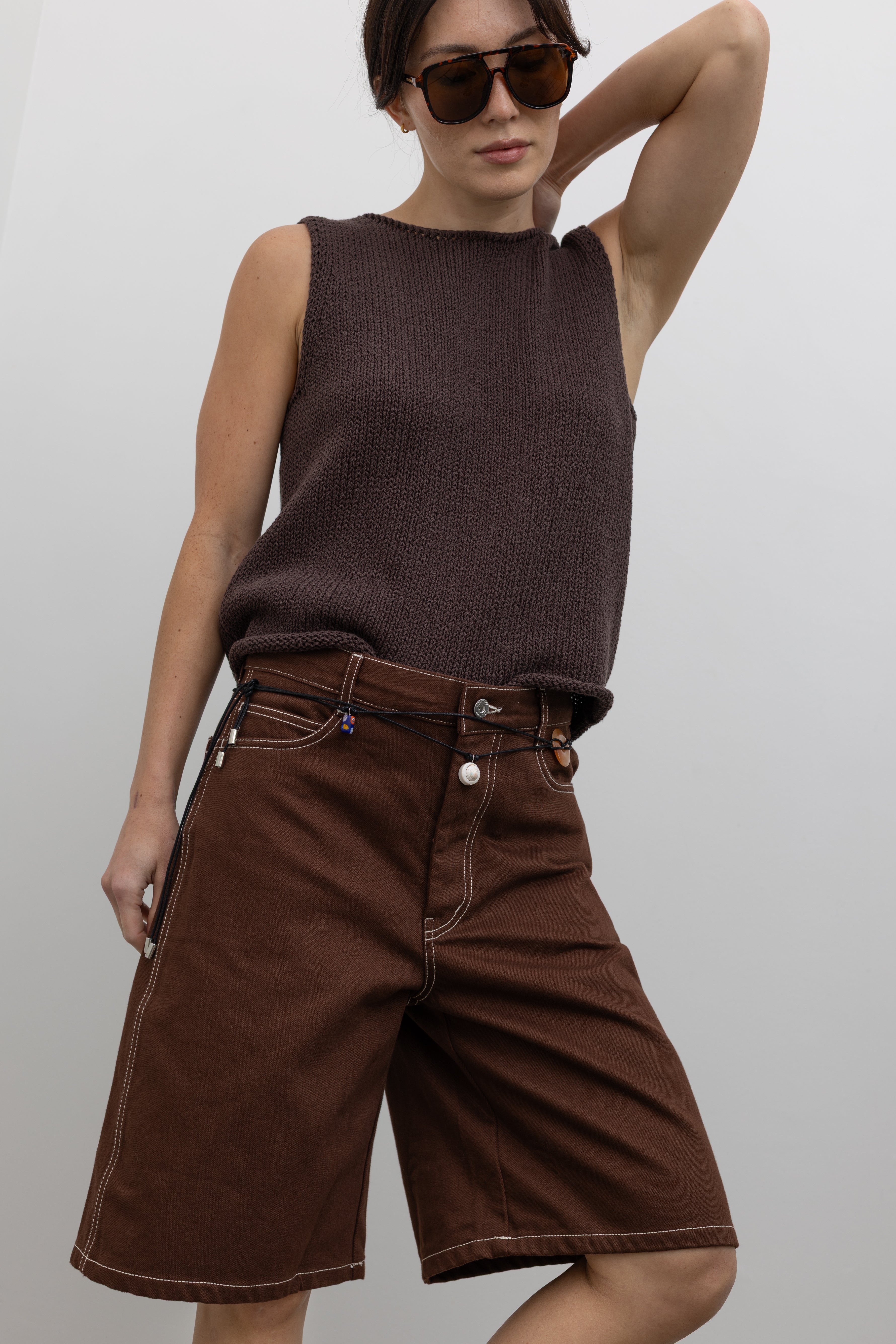 Front view of a woman posing wearing modern minimalist spring and summer fashion in a Mijeong Park dark brown boat neck knit tank featuring a relaxed fit and rolled edges at the neck and hem paired with Kowtow Framework Organic Cotton Brown Denim Long Shorts that sit just above the knee and feature white contrast stitching at an Alas Eius store.  