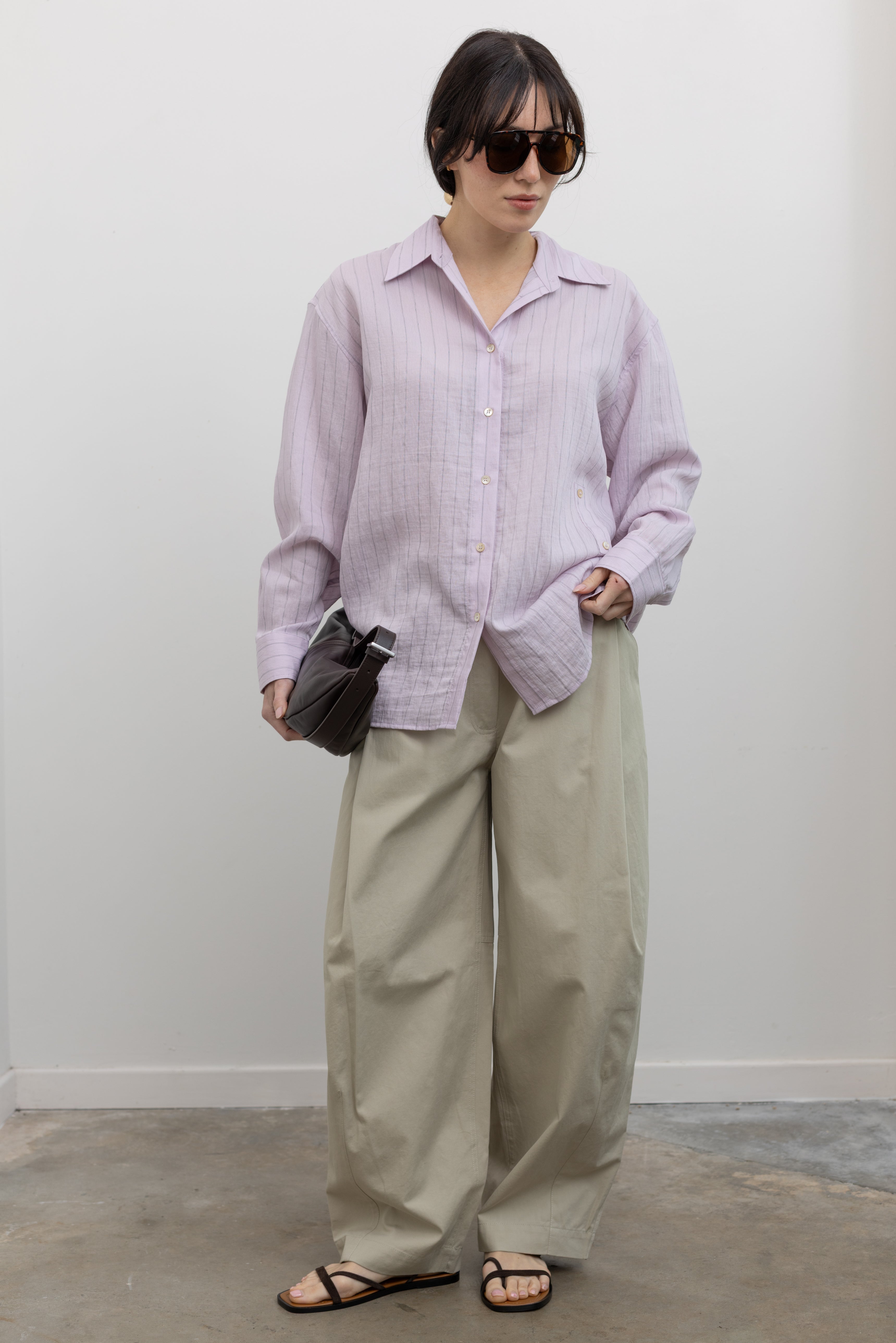 Full length front view of a woman wearing elevated weekend and travel capsule wardrobe staples in a Mijeong Park oversized soft pink pinstripe button down shirt featuring a classic collar, long sleeves, and an off center button down front paired with an elevated classic khaki baggy curved leg pant with strappy sandals at an Alas Eius store. 